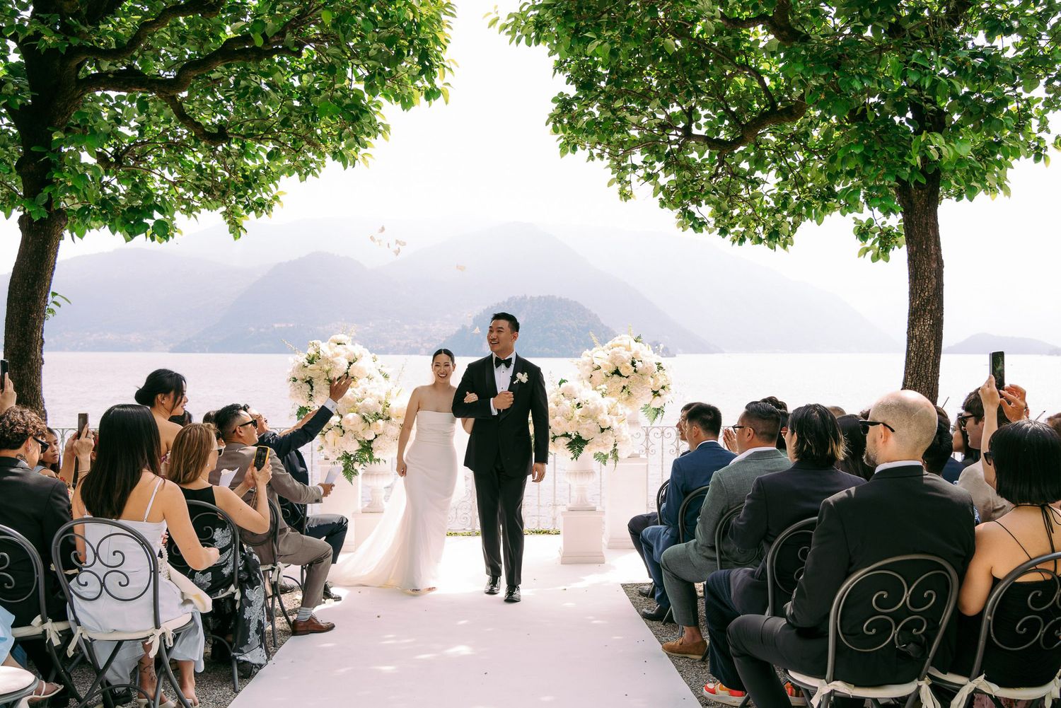 Outdoor wedding ceremony with guests seated in white chairs surrounded by lush green trees overlooking a scenic landscape.