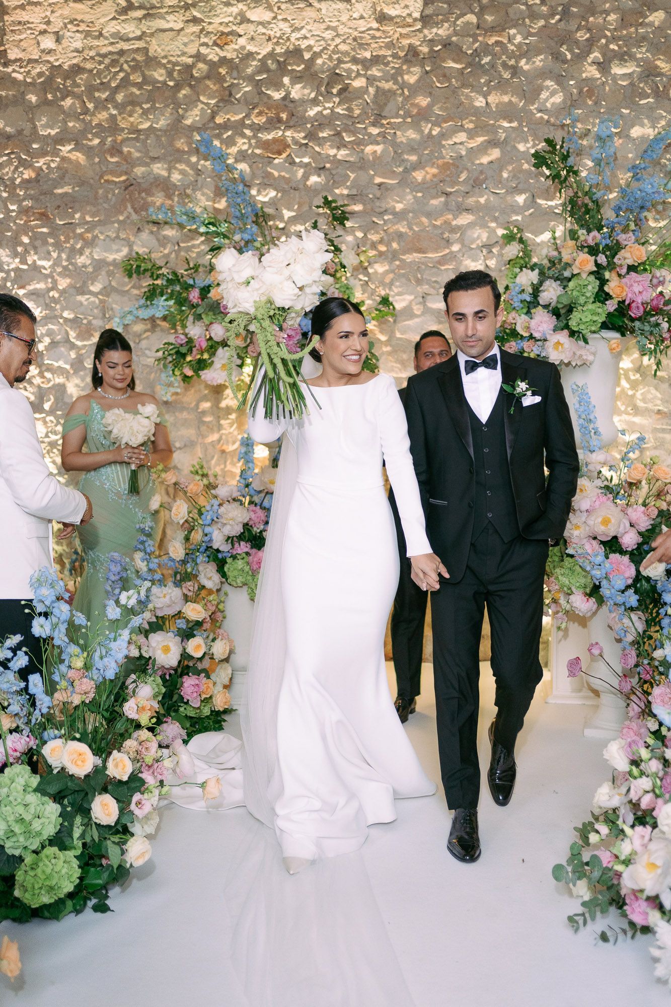 Wedding party walking under a floral archway with pastel pink and blue flowers against a stone wall.