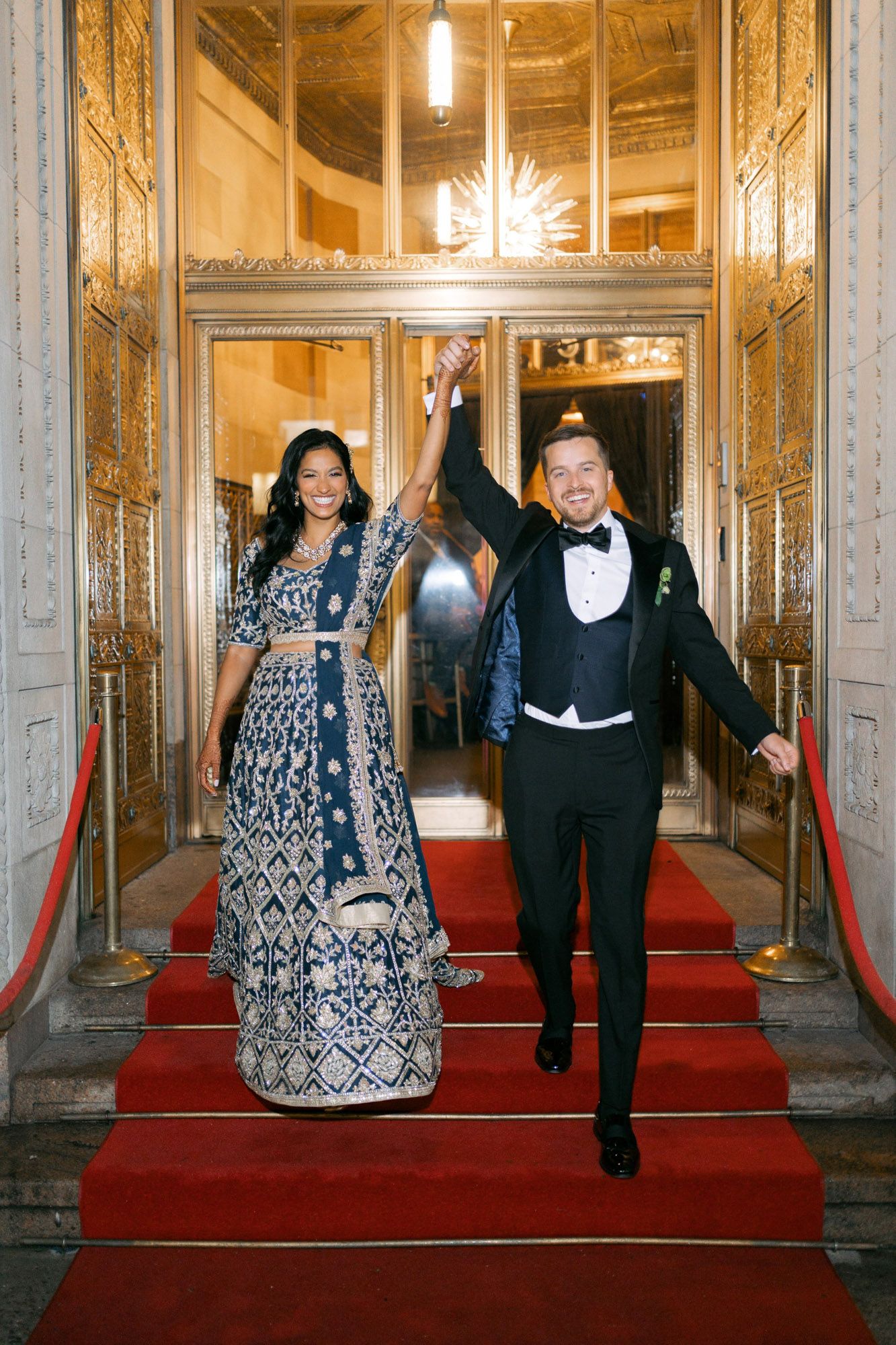 Elegantly dressed guests walk down a grand red-carpeted hallway with ornate gold walls and hanging chandeliers.