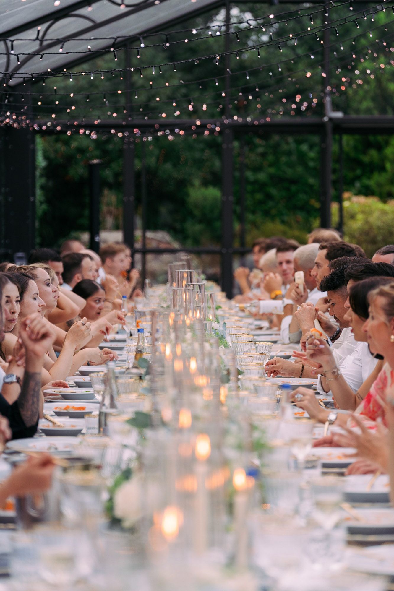 Long outdoor dining table with glowing candles and string lights creating a romantic atmosphere at dusk.