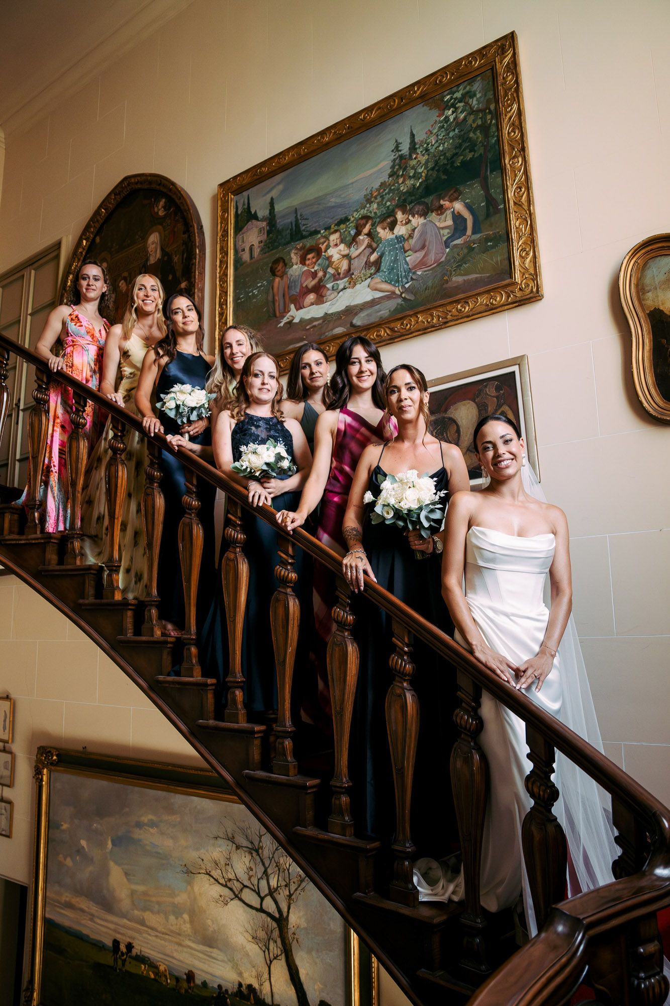 A wedding party poses on an elegant staircase with ornate paintings on the wall.