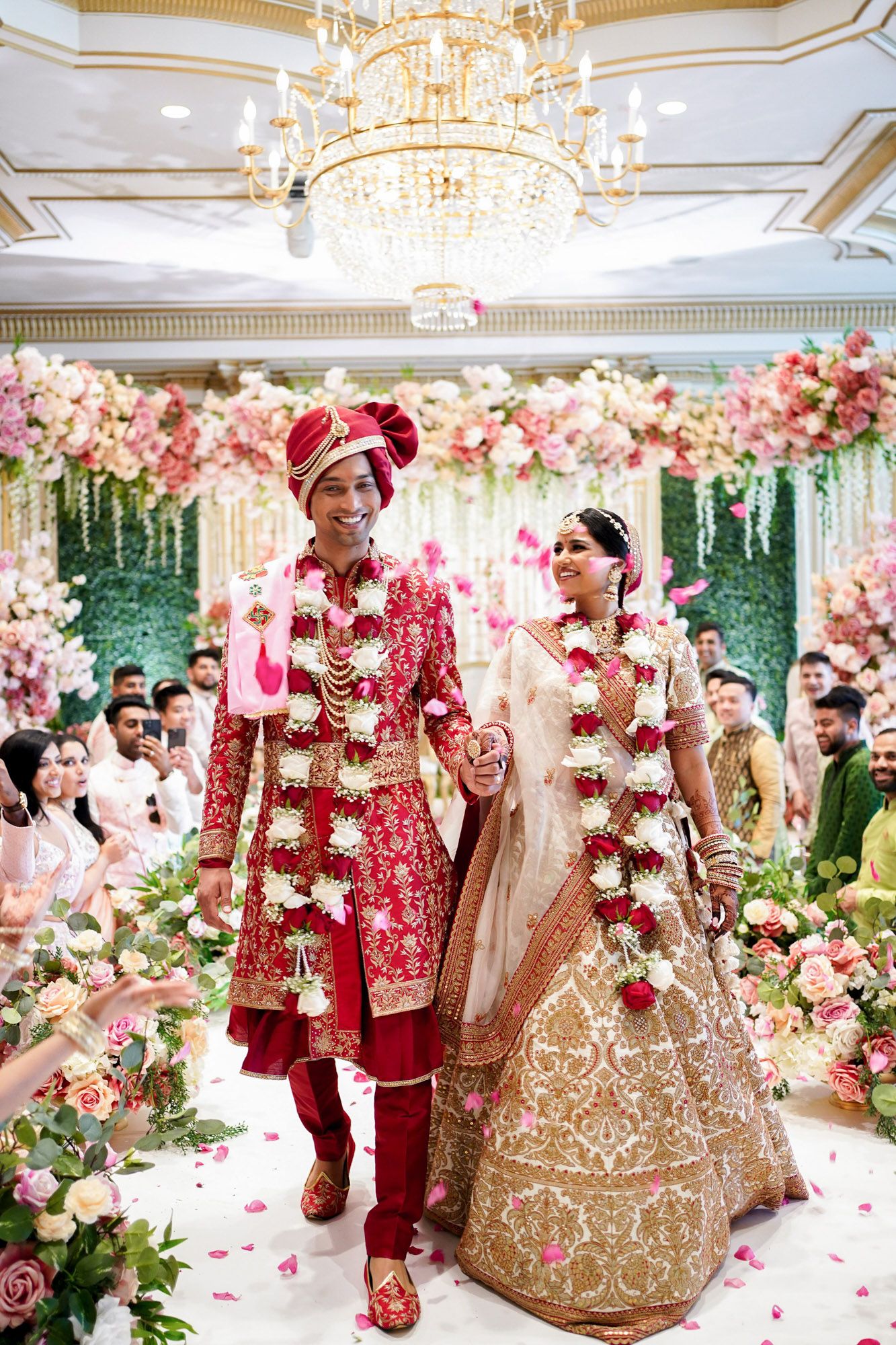 Elegant Indian wedding ceremony with red and gold traditional attire surrounded by pink and white floral decorations.