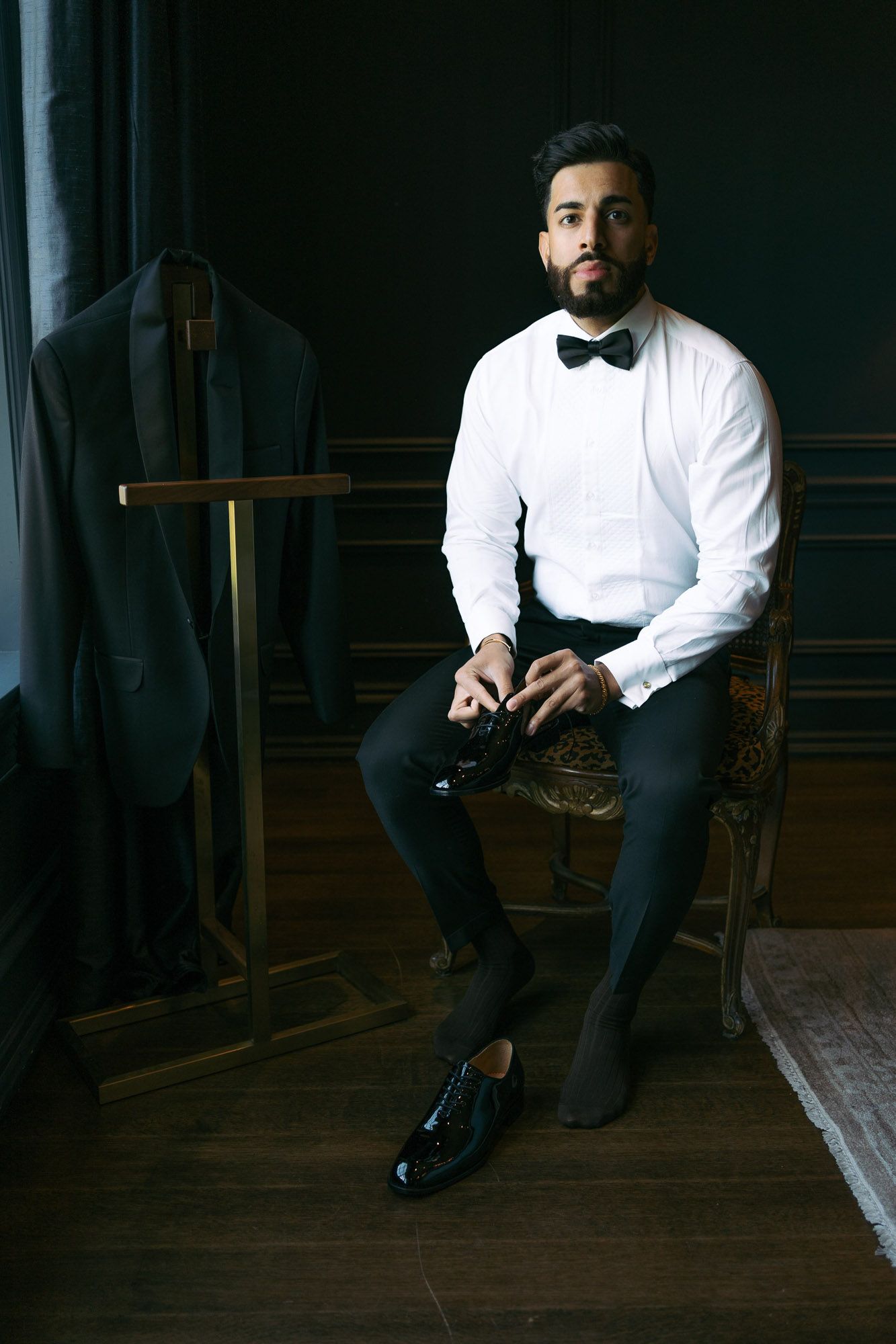 A person in formal attire sits in a dark room wearing a white dress shirt and black bow tie.