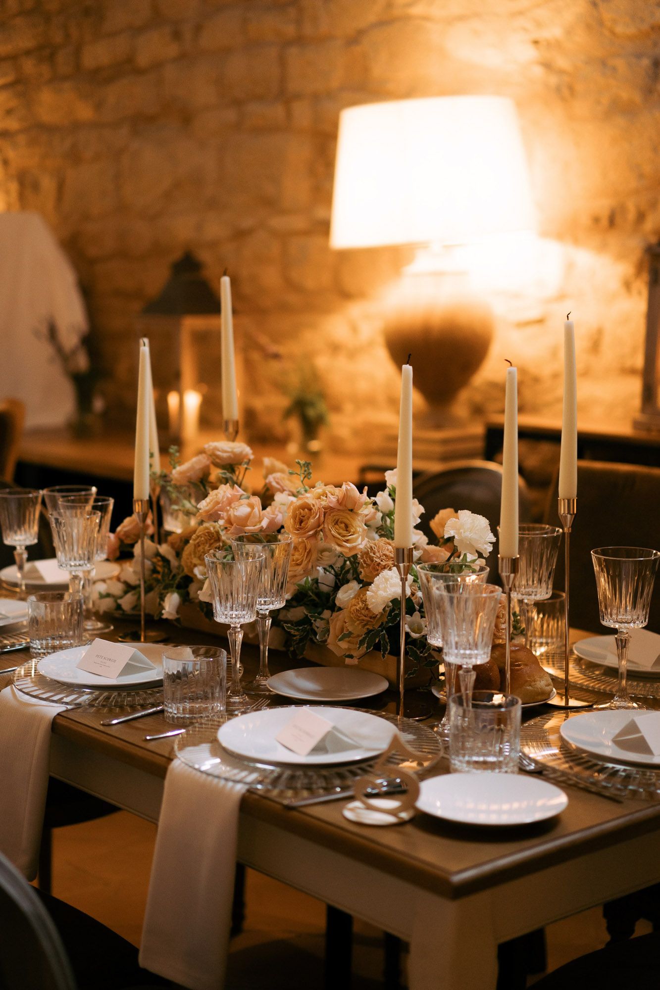 Elegant table setting with glowing candlelight illuminating white plates and crystal glasses in a rustic stone venue.