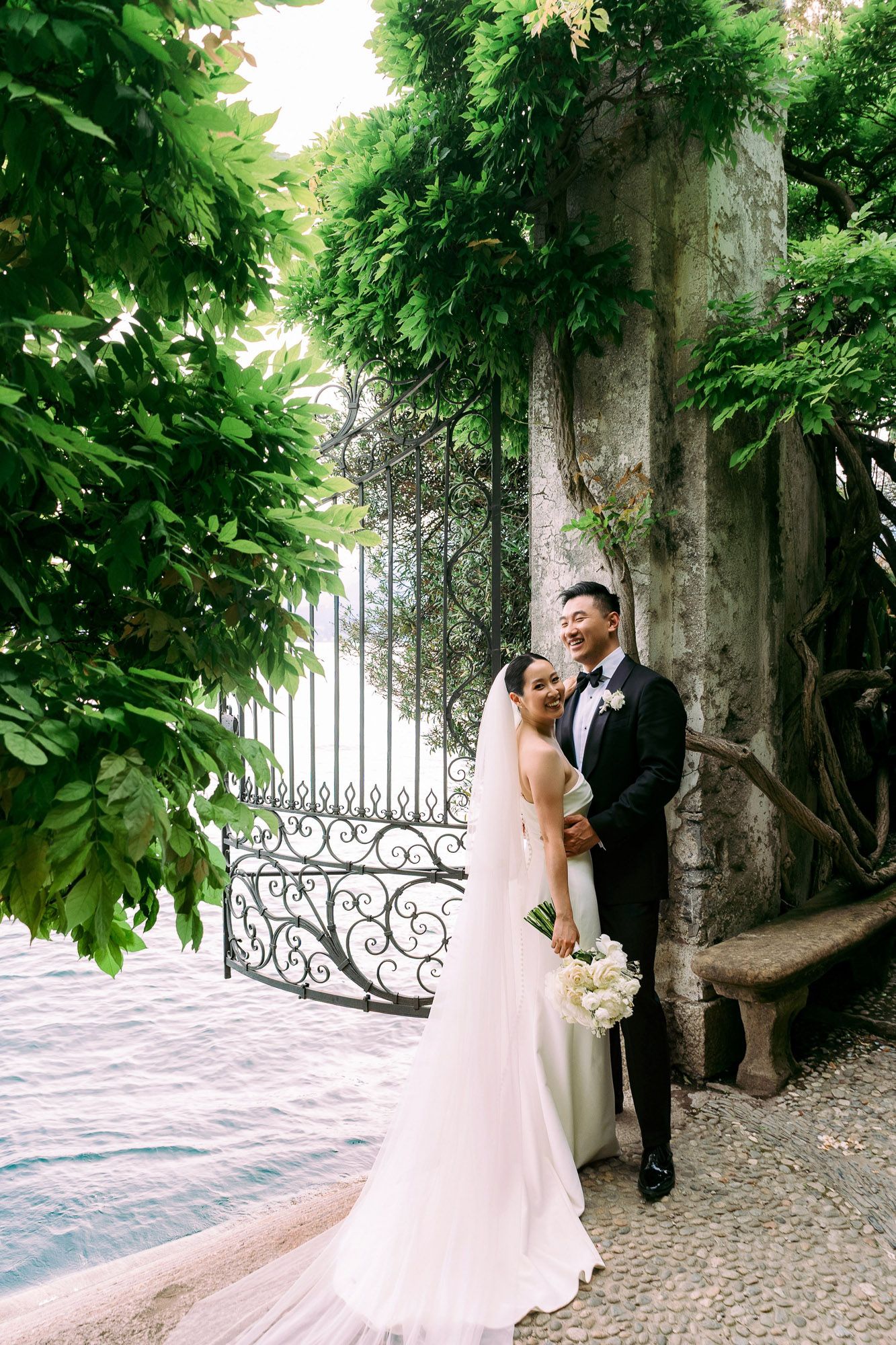 A couple in wedding attire poses by an ornate iron gate and lush greenery in an elegant garden setting.