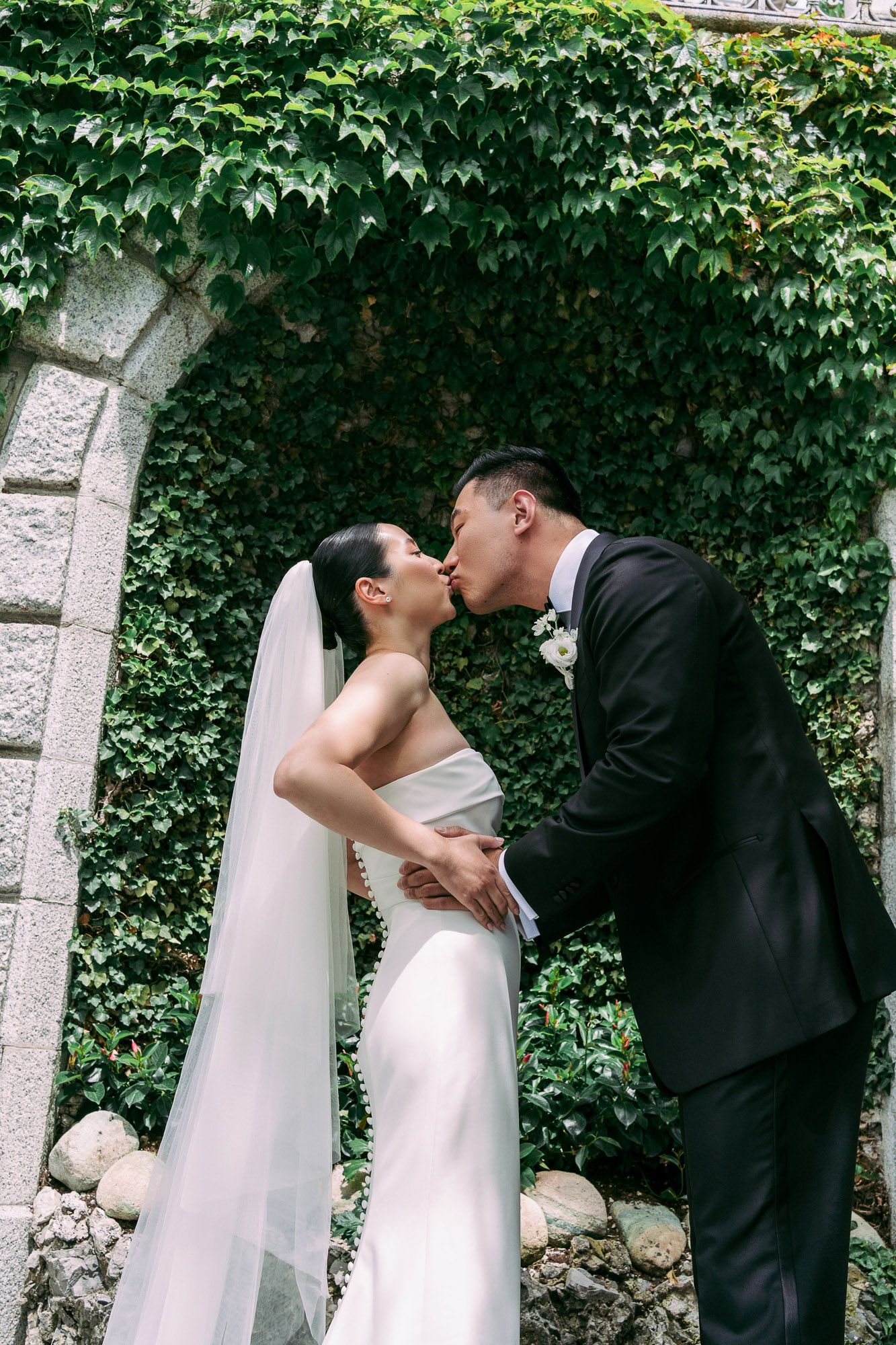 A romantic wedding couple shares a tender kiss in front of an ivy-covered stone wall archway.