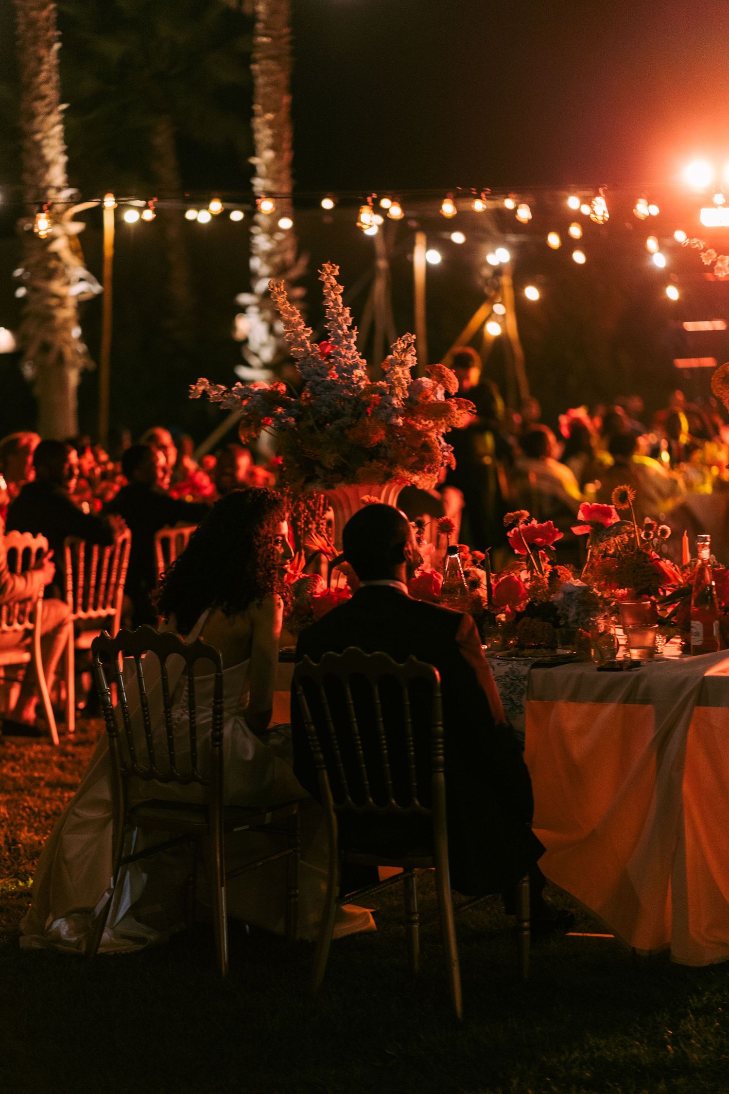 Romantic evening reception with tables illuminated by warm string lights under palm trees.