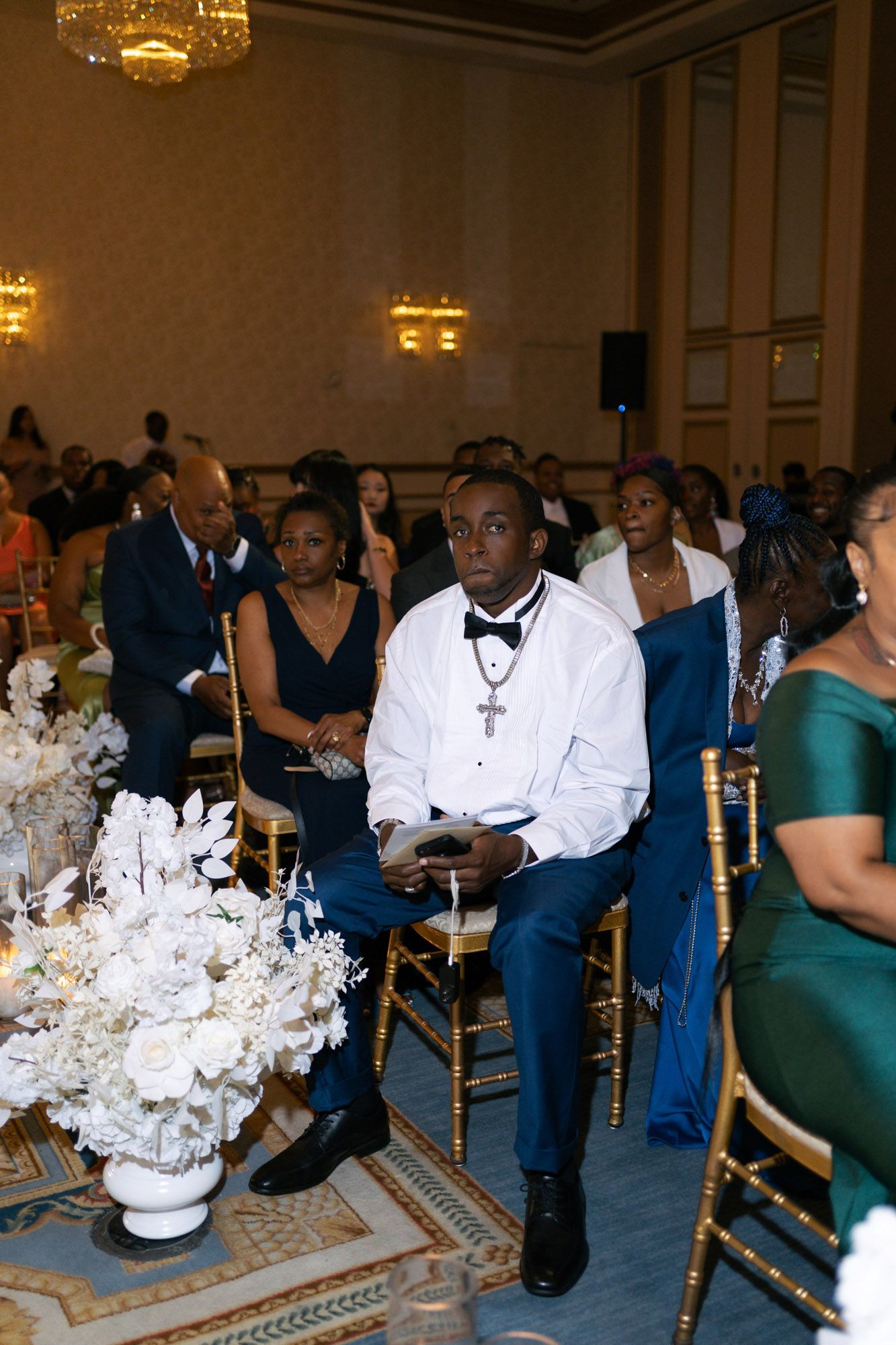 Wedding guests seated at elegant reception tables with white floral centerpieces and gold chairs in formal ballroom.