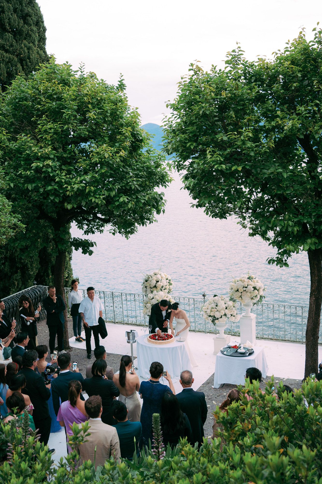 Outdoor wedding ceremony overlooking a lake with floral arrangements and guests gathering under lush green trees.