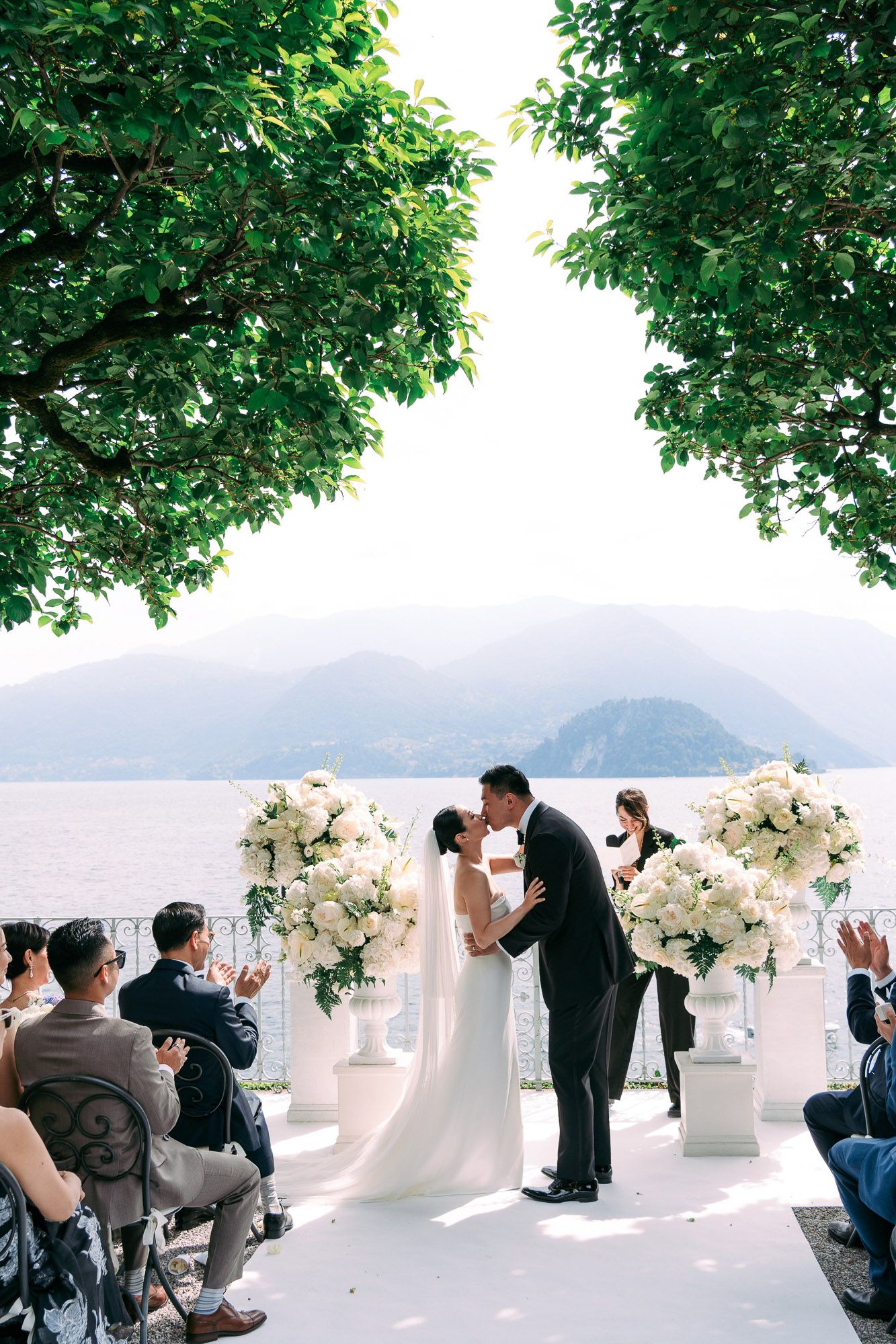 Wedding photographers capturing a lakeside ceremony from various angles with white floral arrangements and a mountain backdrop.