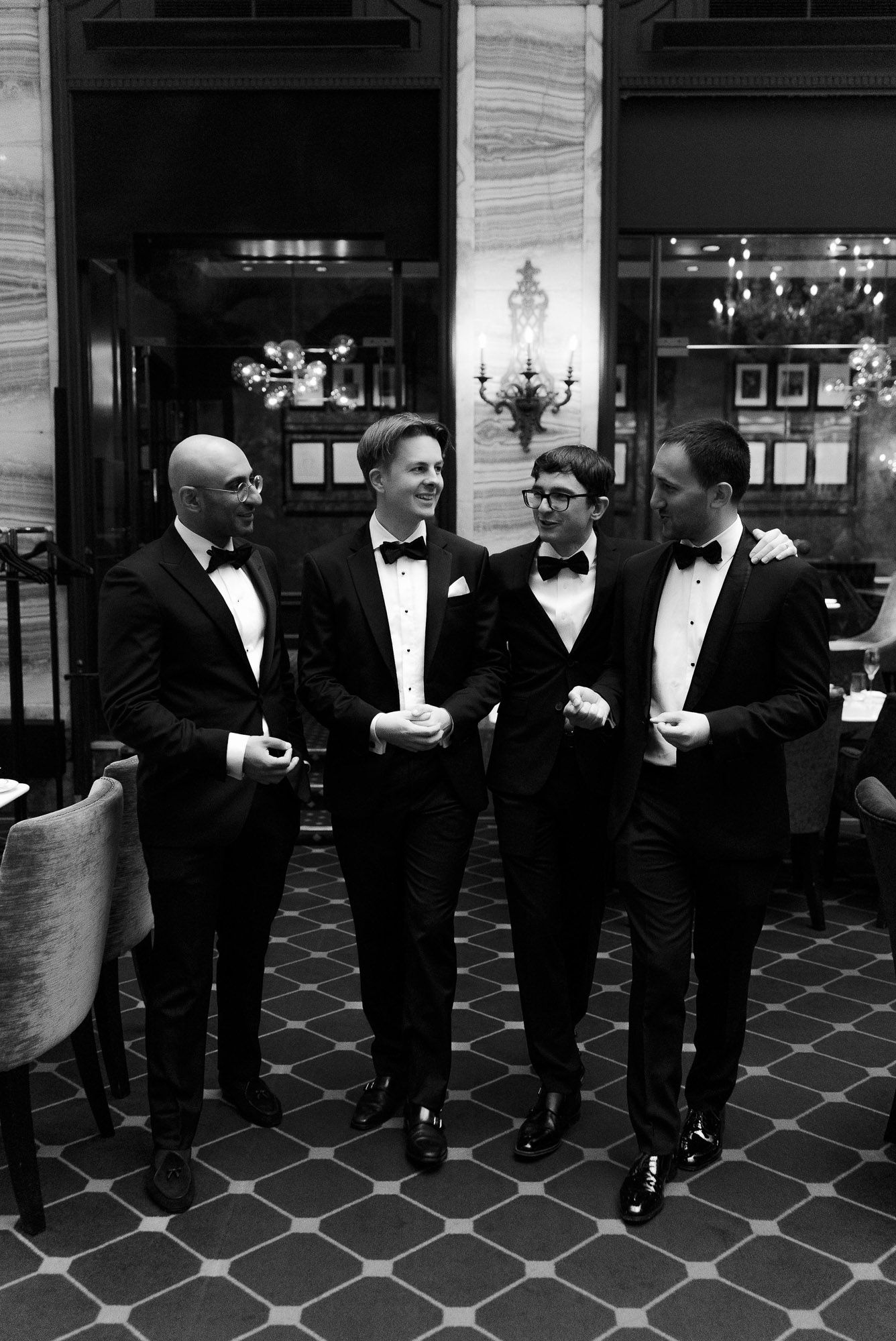 A group of men in formal black tuxedos and bow ties stand in a line against a tiled wall at an event.