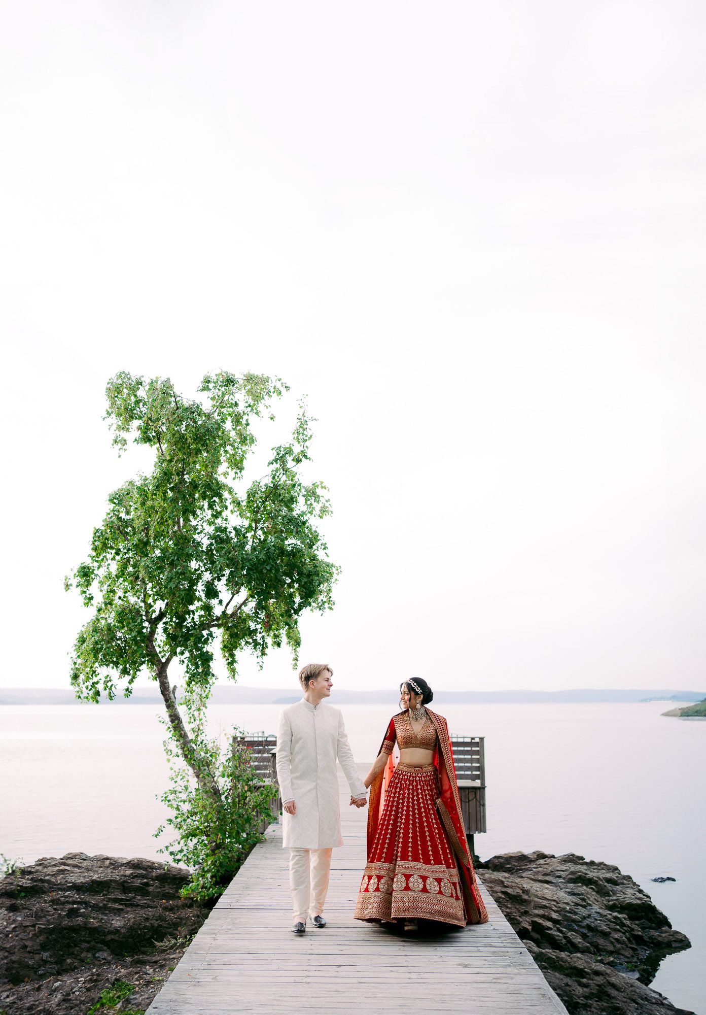 Couple in traditional Indian wedding attire stands together on rocky lakeside shore with tree and dock in background.
