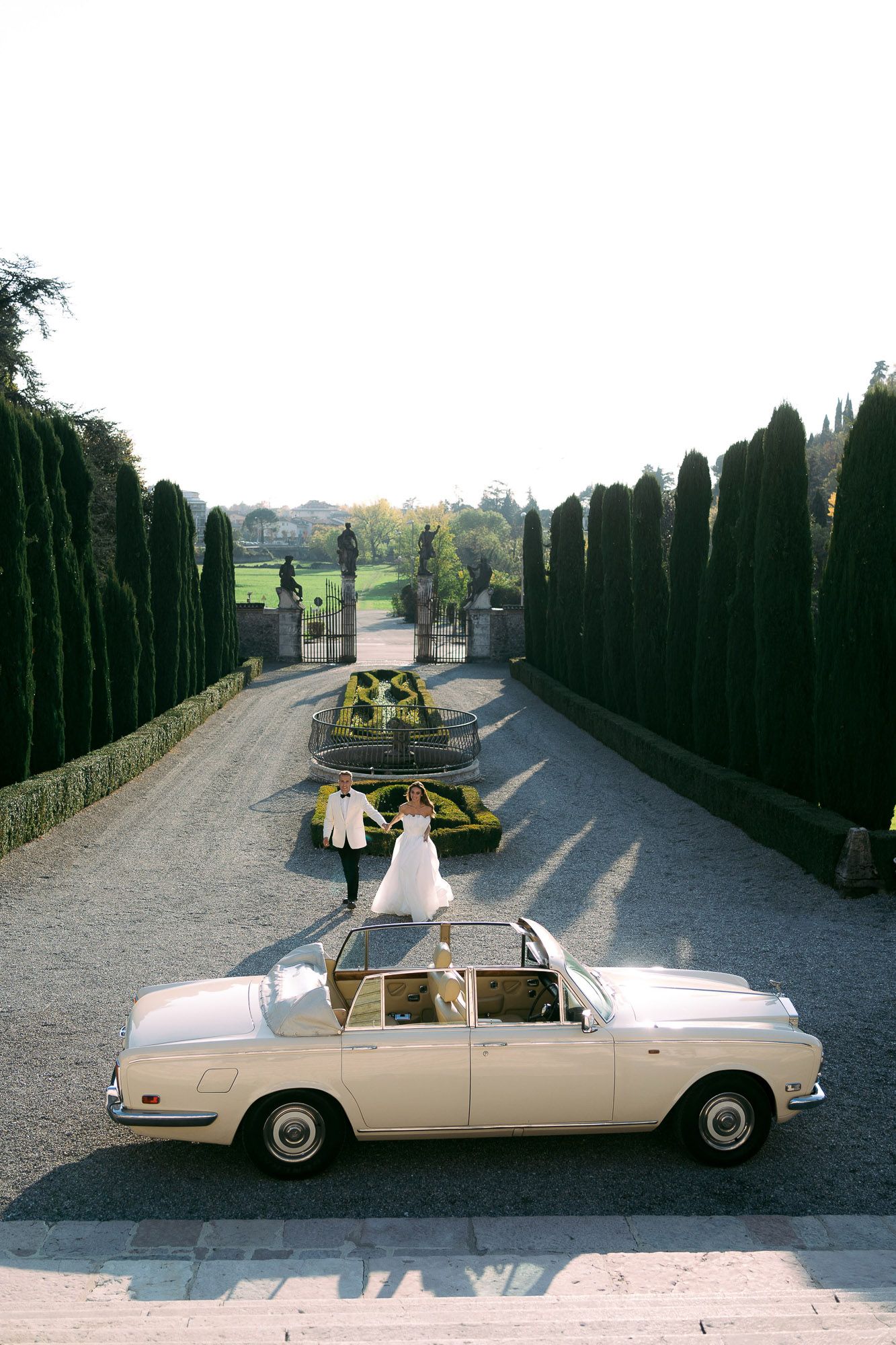 Classic cream-colored luxury car decorated with yellow flowers and greenery for a wedding procession on a tree-lined path.