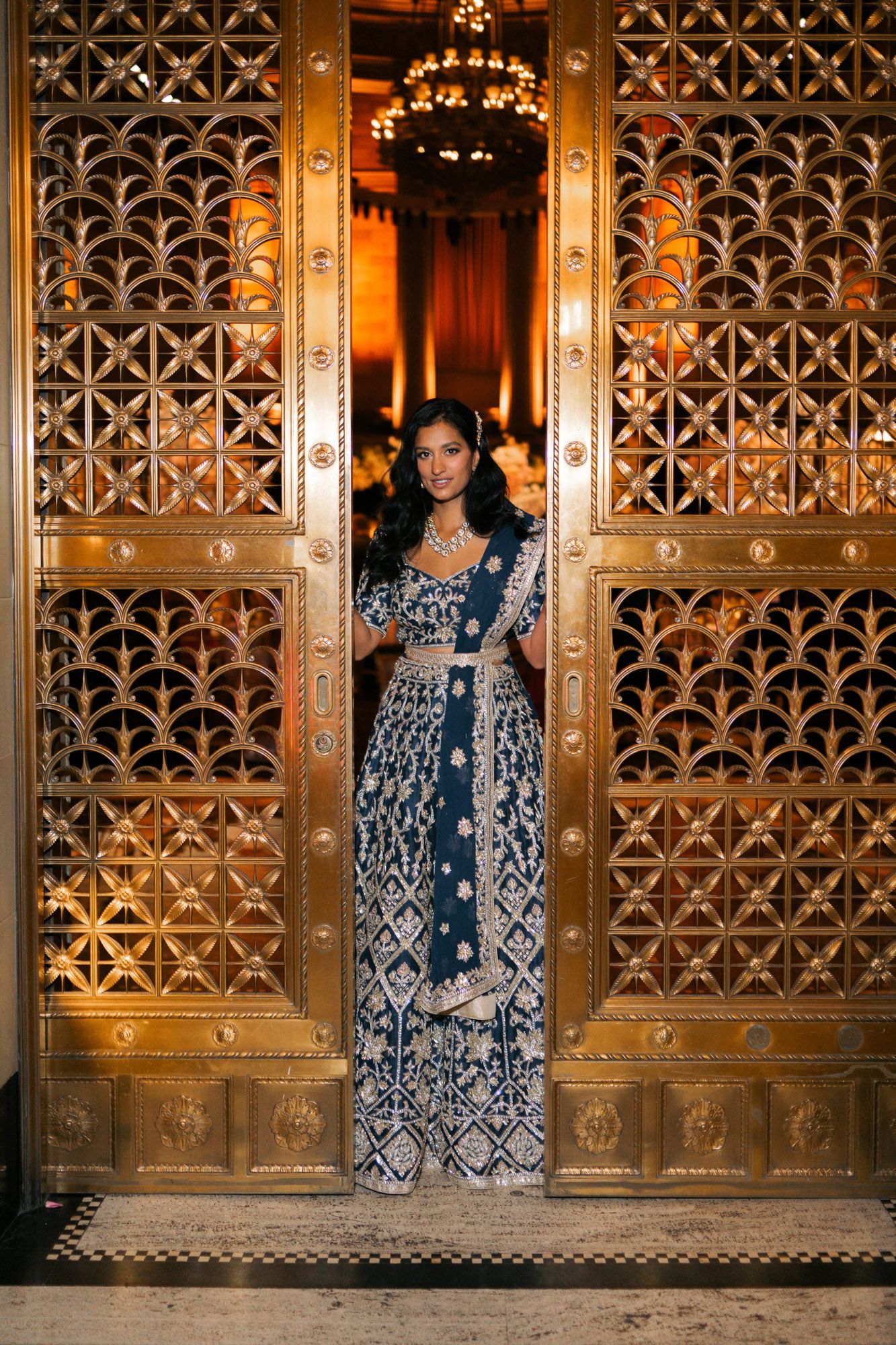 A person in an ornate navy blue and white patterned dress poses against decorative gold metalwork doors.