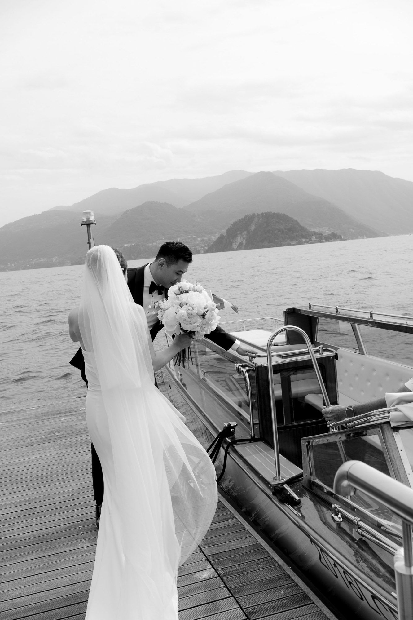 A couple in wedding attire shares a romantic moment on a wooden dock overlooking a scenic mountain lake.