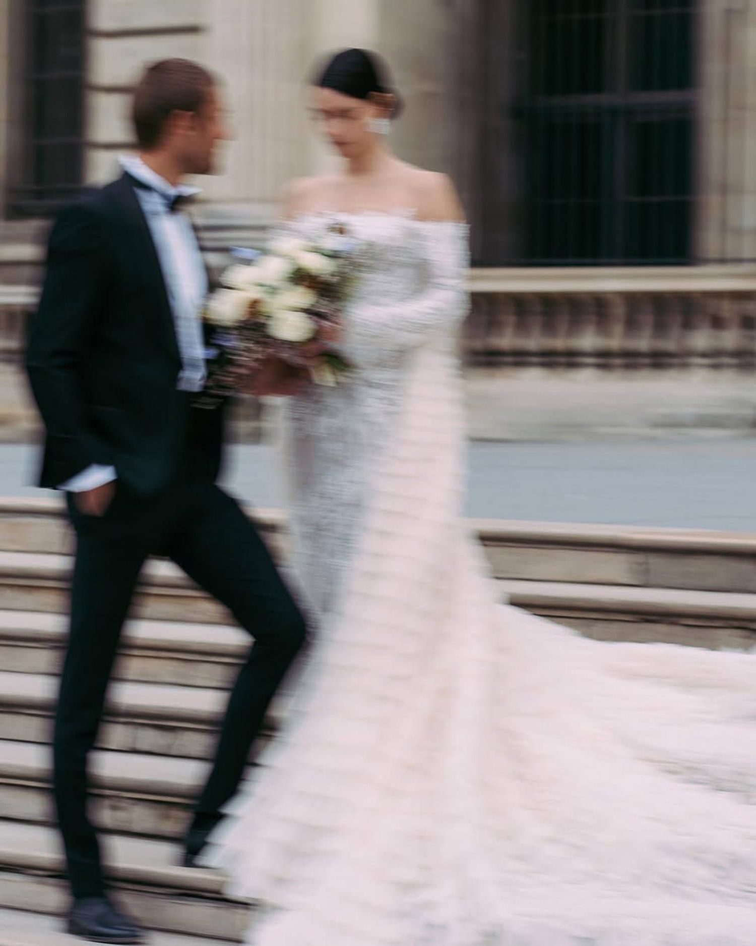 Motion blur captures a bride and groom walking together in formal wedding attire on a city street.