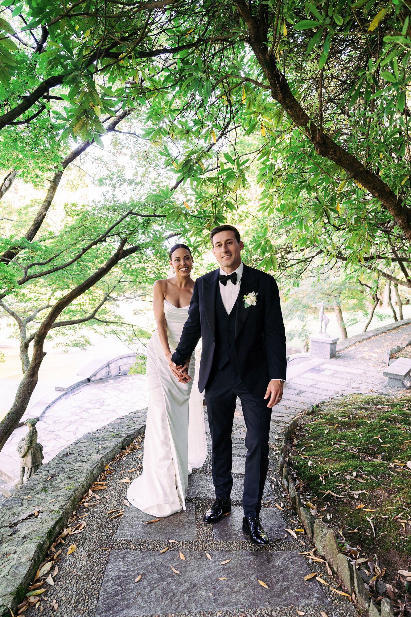 A couple in formal wedding attire poses together along a scenic tree-lined path with natural stone features.