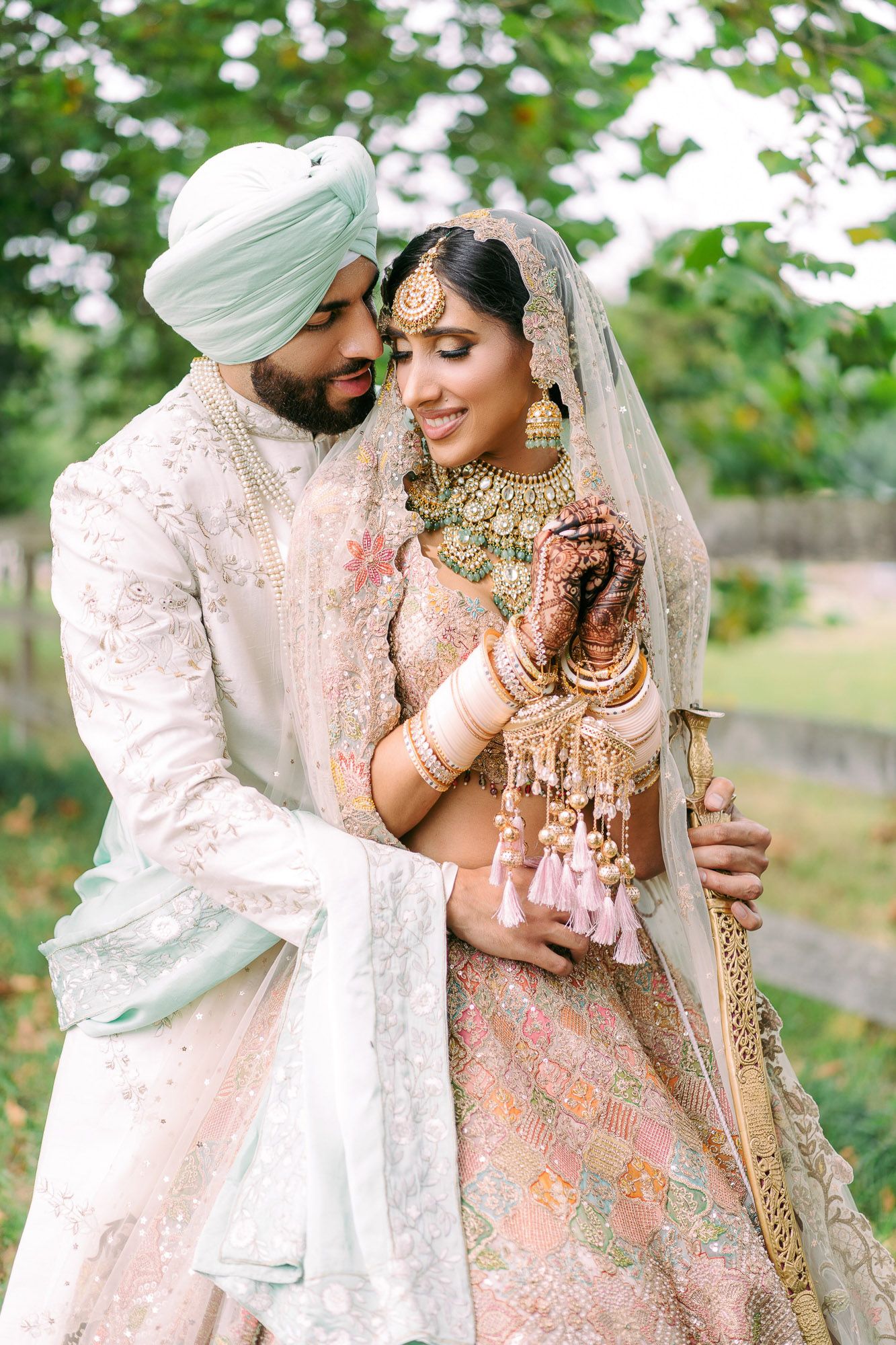Elegant Sikh wedding couple embracing in garden setting wearing traditional white and mint ceremonial attire.