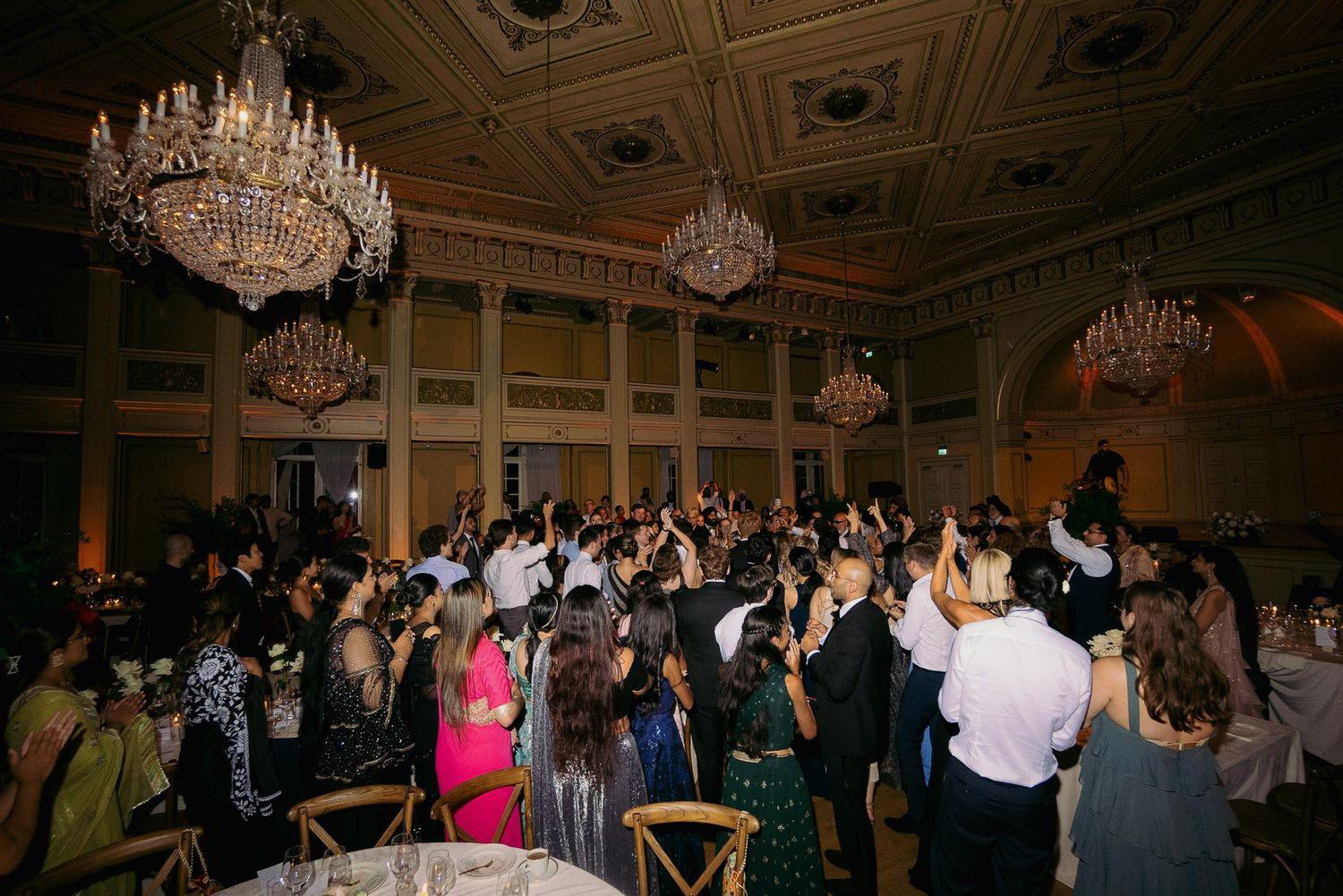 Wedding guests dance and celebrate on an elegant ballroom floor under ornate crystal chandeliers.