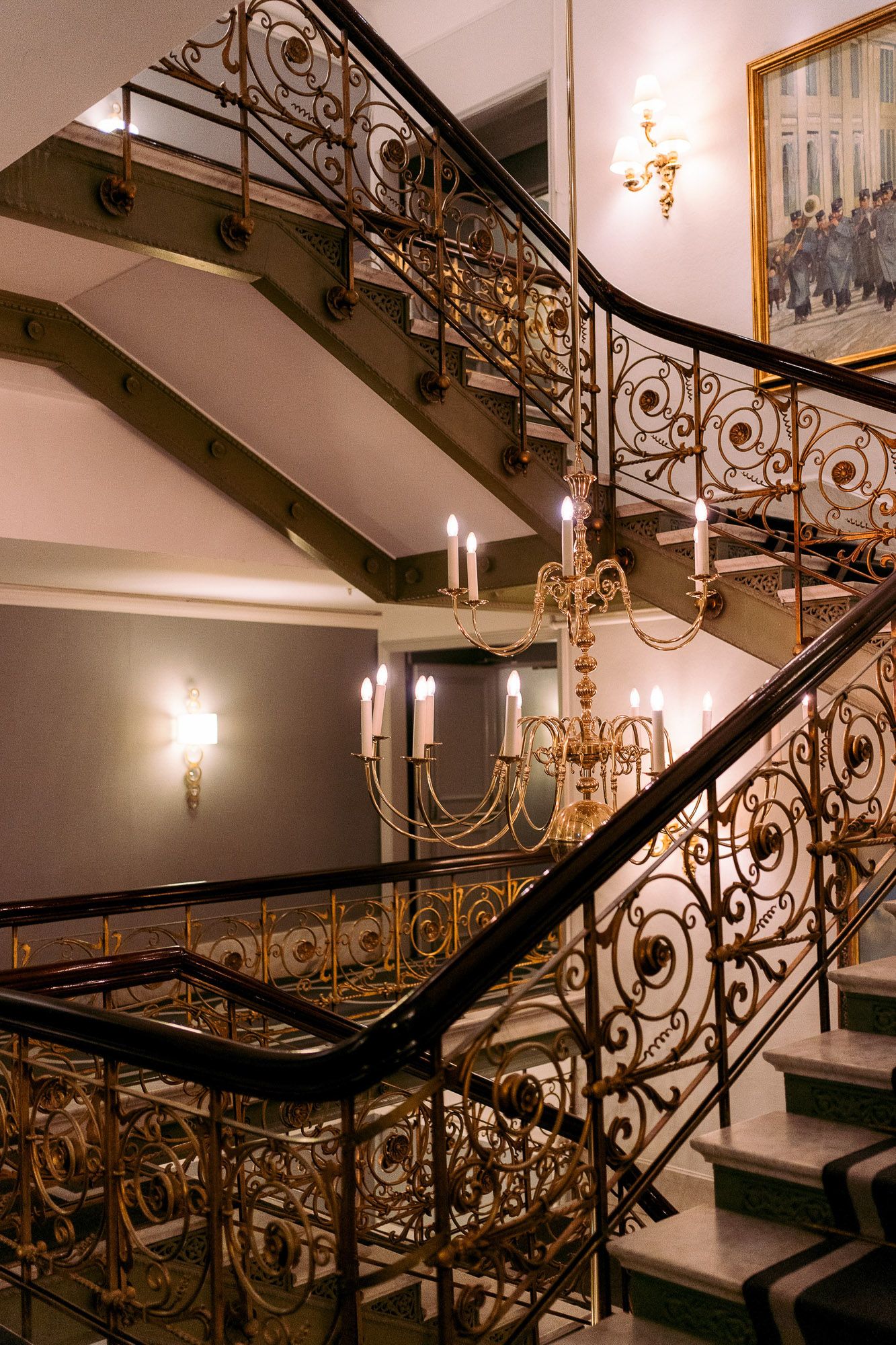 Elegant curved staircase with ornate gold wrought iron railings and a brass chandelier in a luxury interior setting.