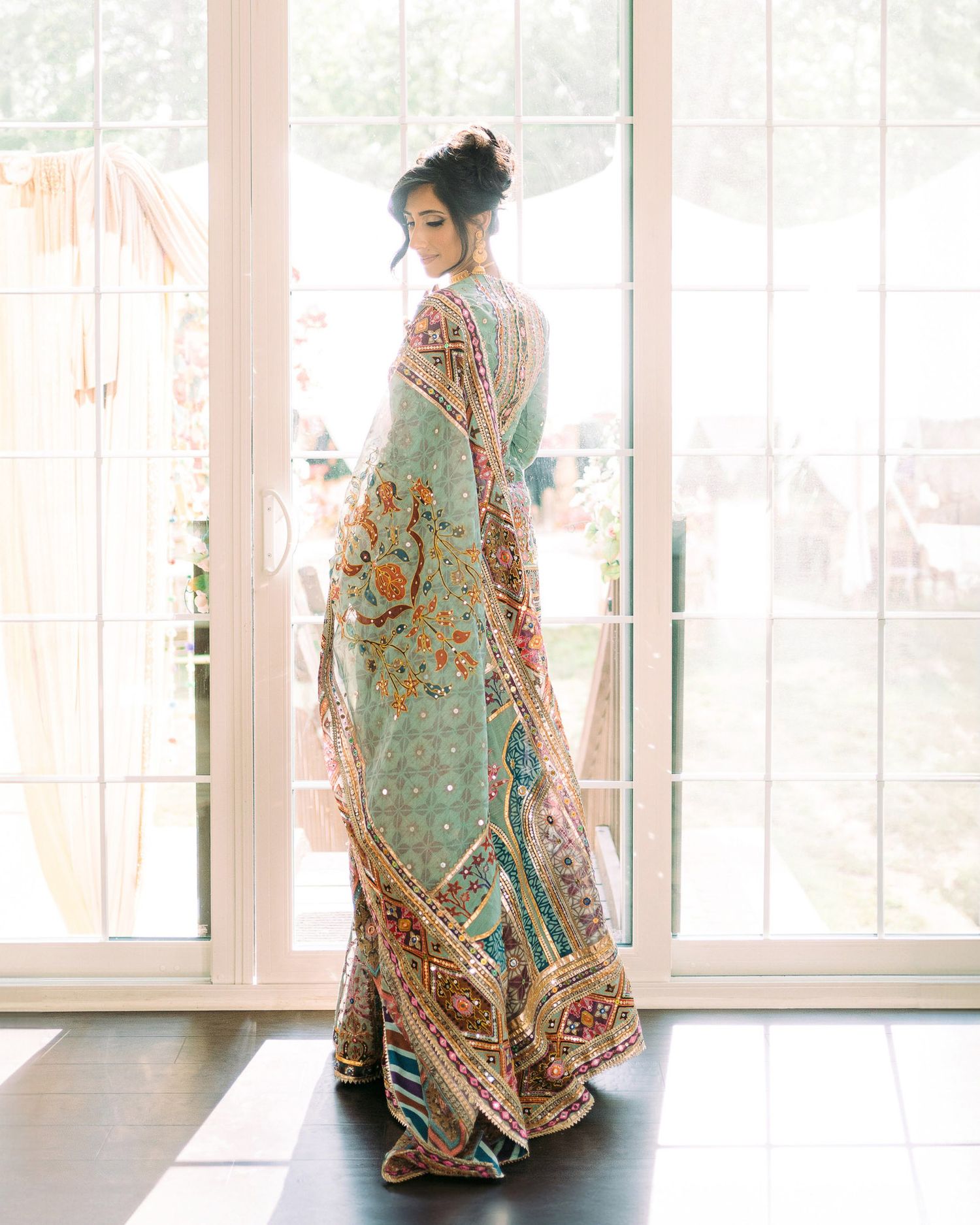 A model poses in an elegant mint green floral embroidered Indian bridal gown in a bright sunlit room.