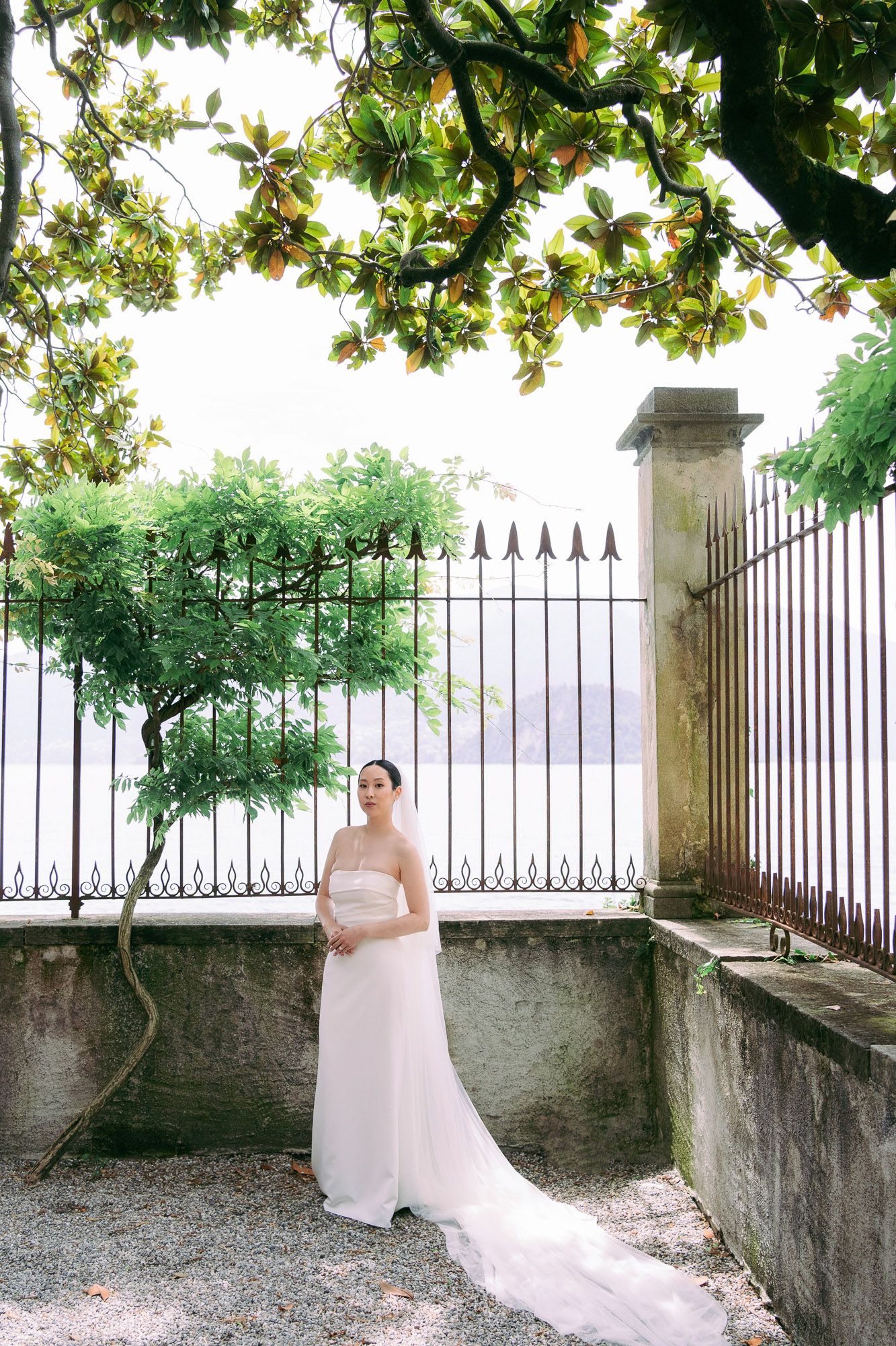 A bride in a flowing white dress poses in a garden setting framed by ornate iron gates and lush greenery.