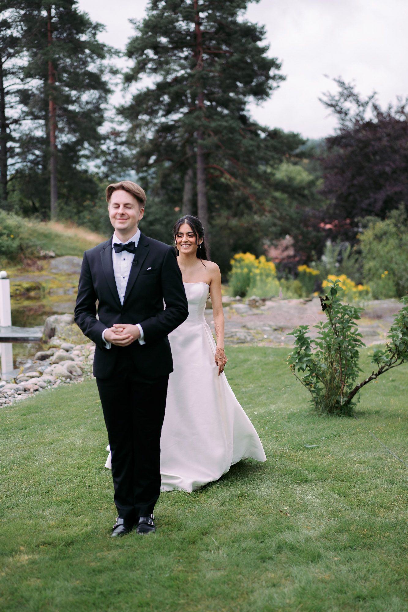A couple in wedding attire walk together on a garden path surrounded by evergreen trees and flowers.