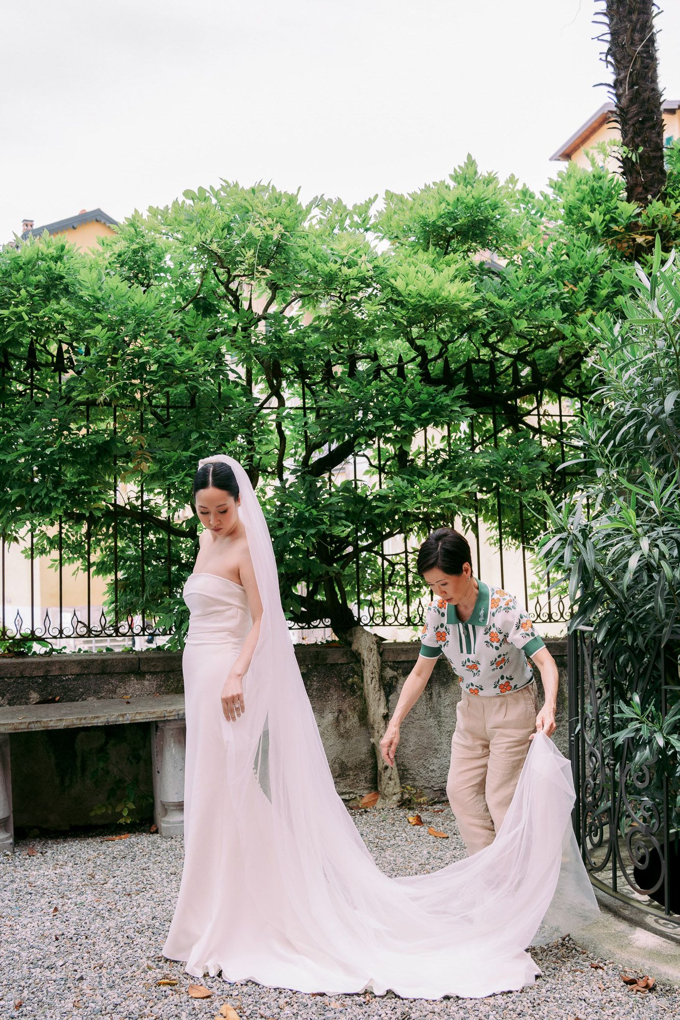 Bride in flowing white dress walking along stone path with trailing veil in garden setting.