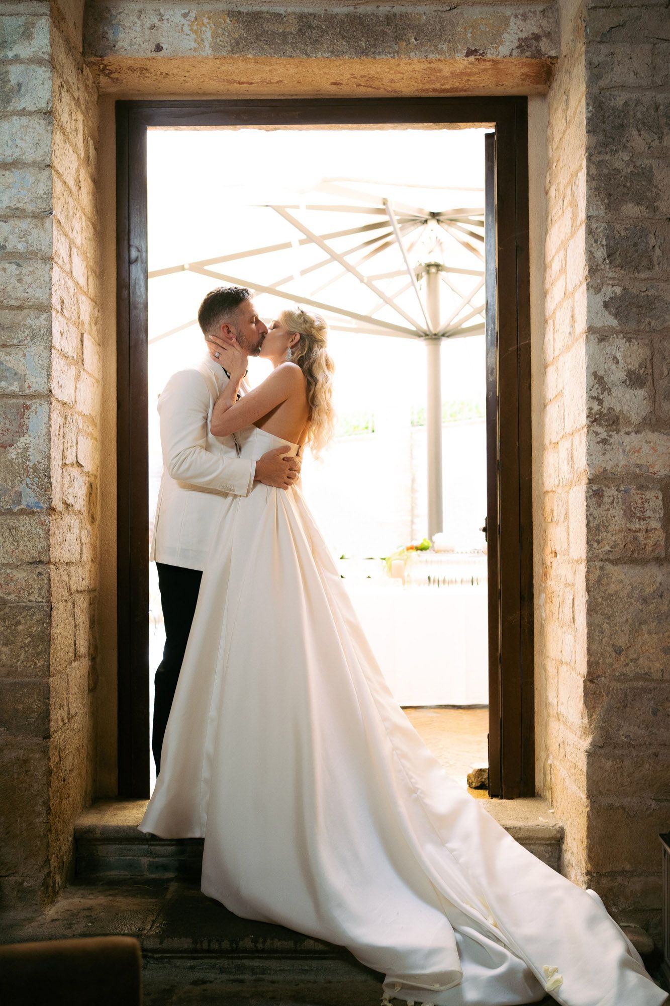 A romantic wedding couple shares an intimate moment in a rustic stone building with natural light streaming through windows.