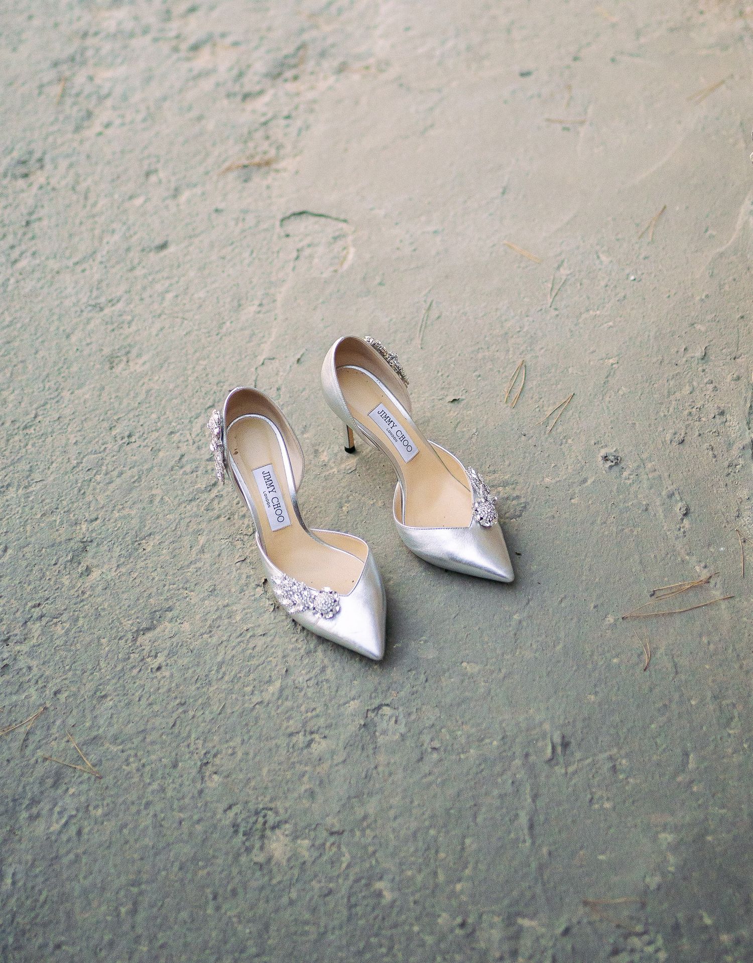 Elegant silver pointed-toe bridal heels with sparkly embellishments sit on a textured grey surface.