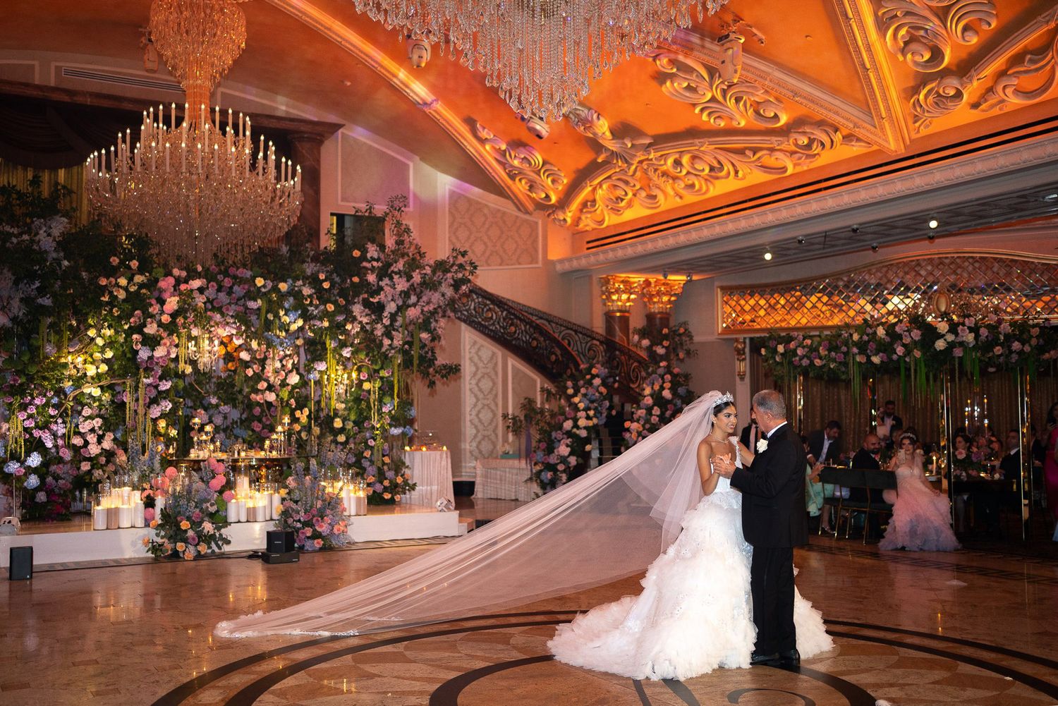 A luxurious ballroom wedding reception venue with ornate chandeliers and floral decorations glowing in warm amber lighting.