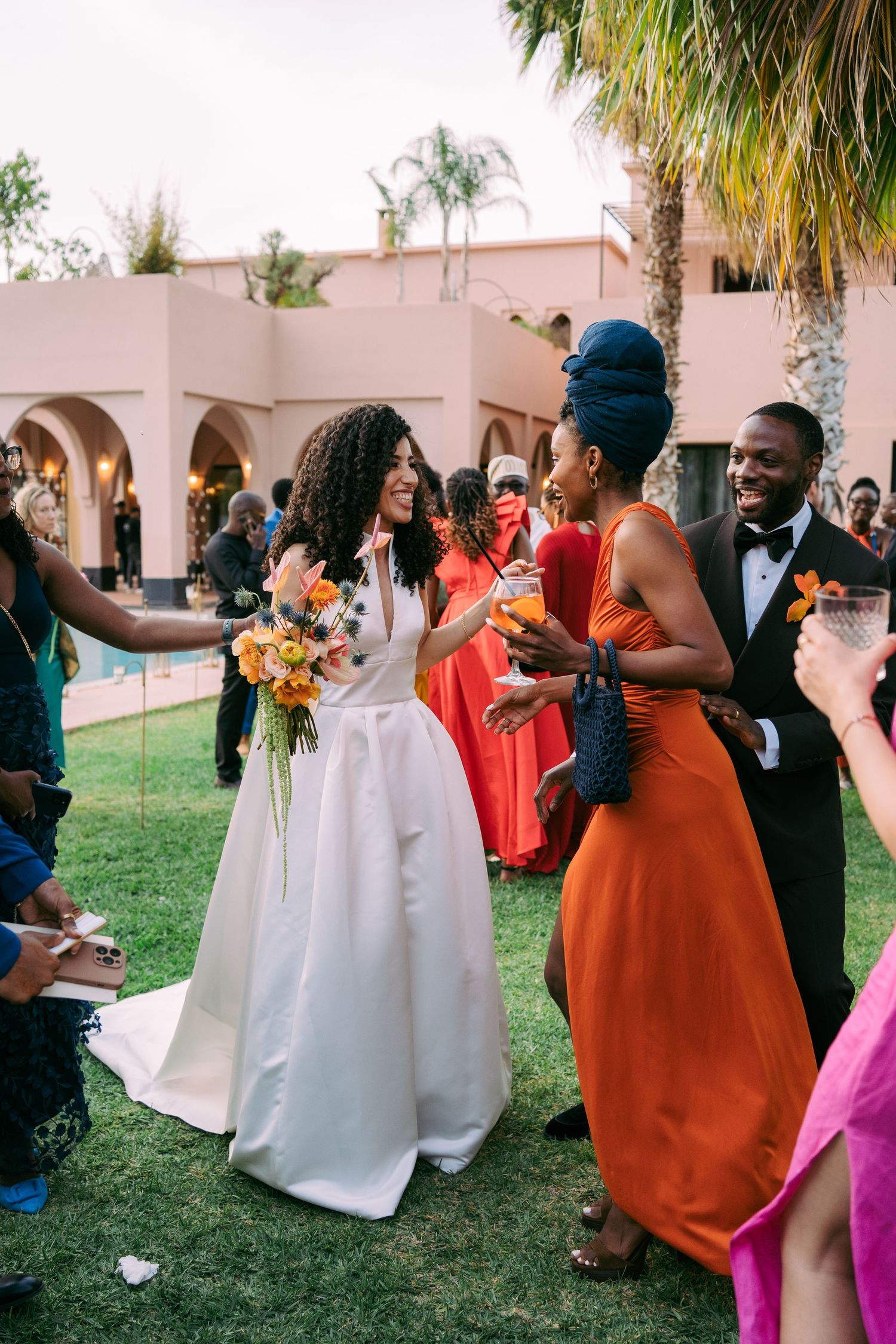 Wedding guests in vibrant attire celebrate on a lawn with Spanish-style architecture in background.