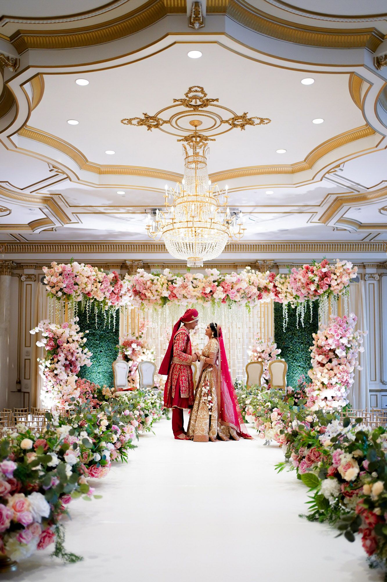 Luxurious wedding venue with ornate gold ceiling, crystal chandeliers and pink floral arrangements along a white aisle.