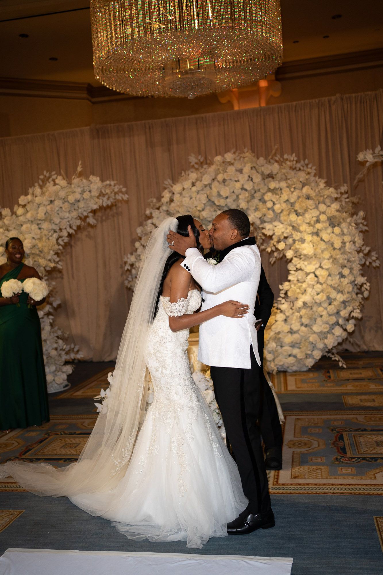 A romantic wedding ceremony moment in front of an elegant white floral arch with a crystal chandelier overhead.