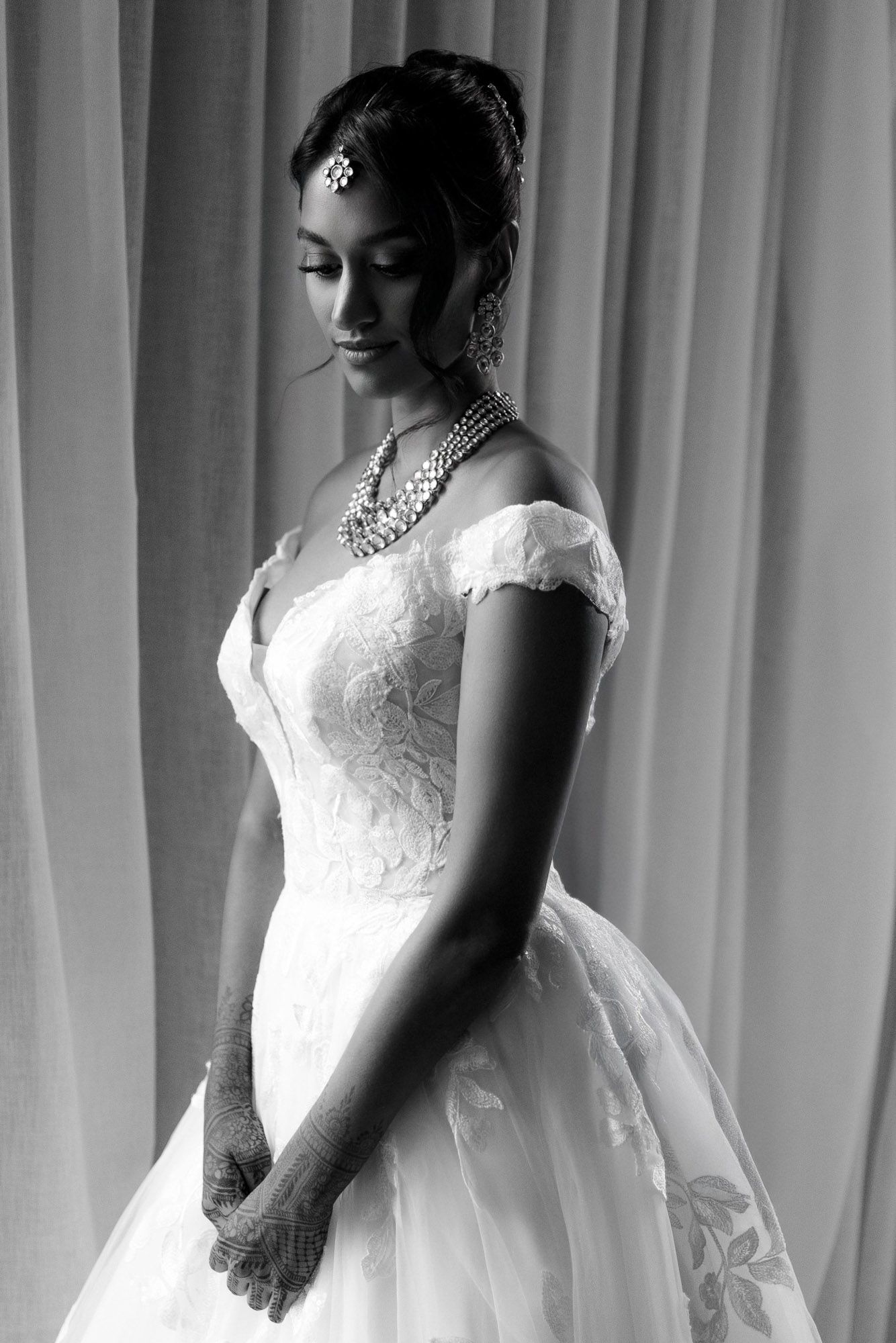Black and white photo of a bride in a lace wedding dress standing against a curtained backdrop.