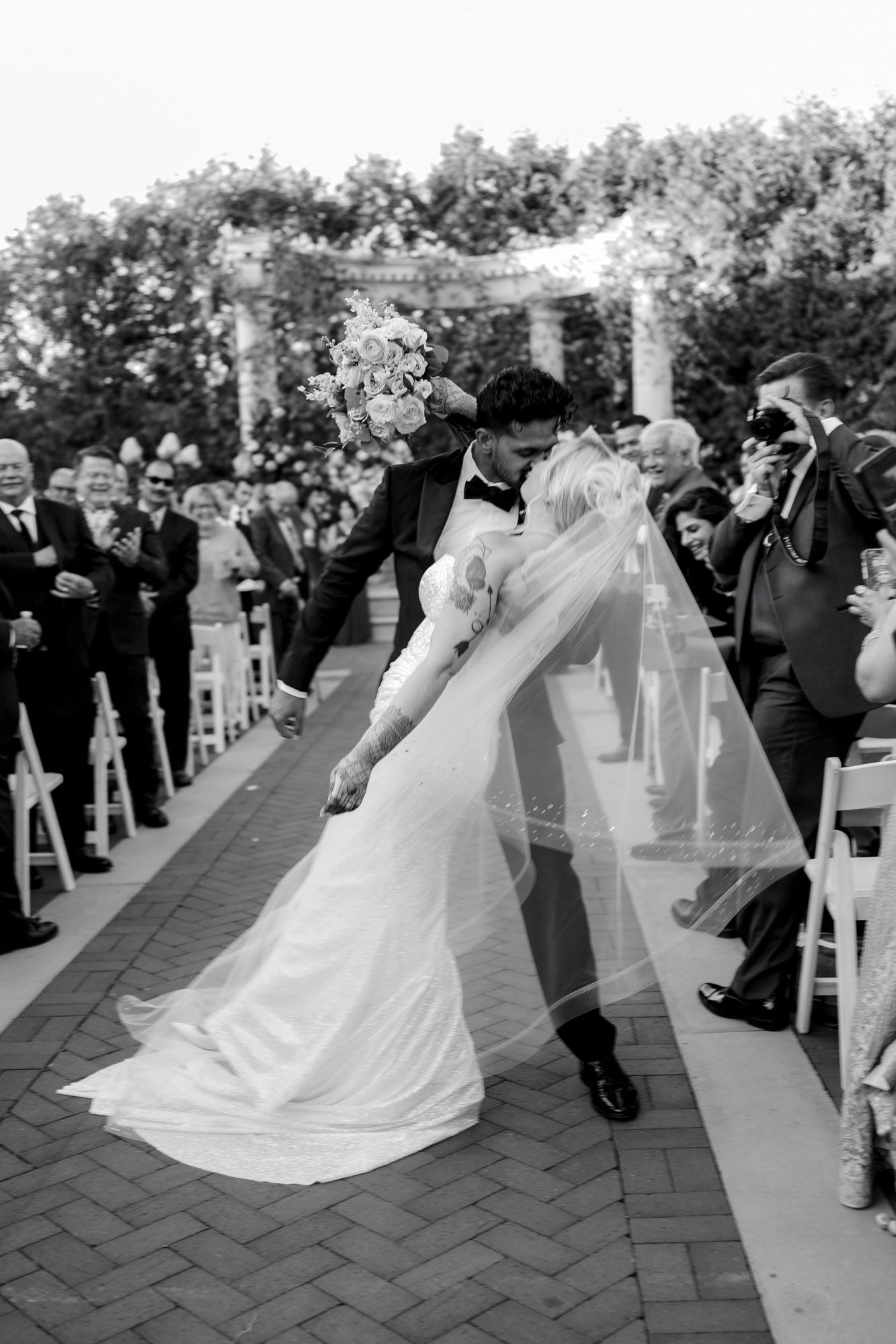 A bride and groom dance together during their outdoor wedding ceremony while guests watch from white chairs.