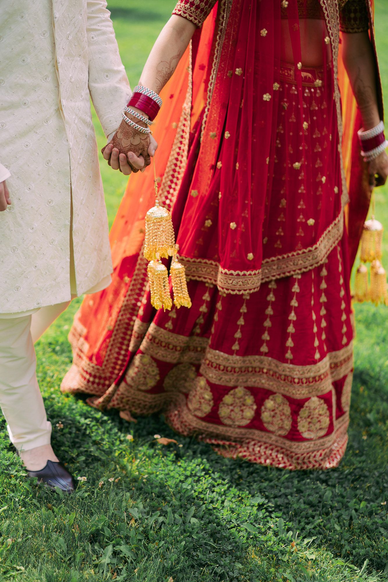 Traditional Indian wedding attire showing red and orange embroidered lehenga with gold details and white sherwani on grass.