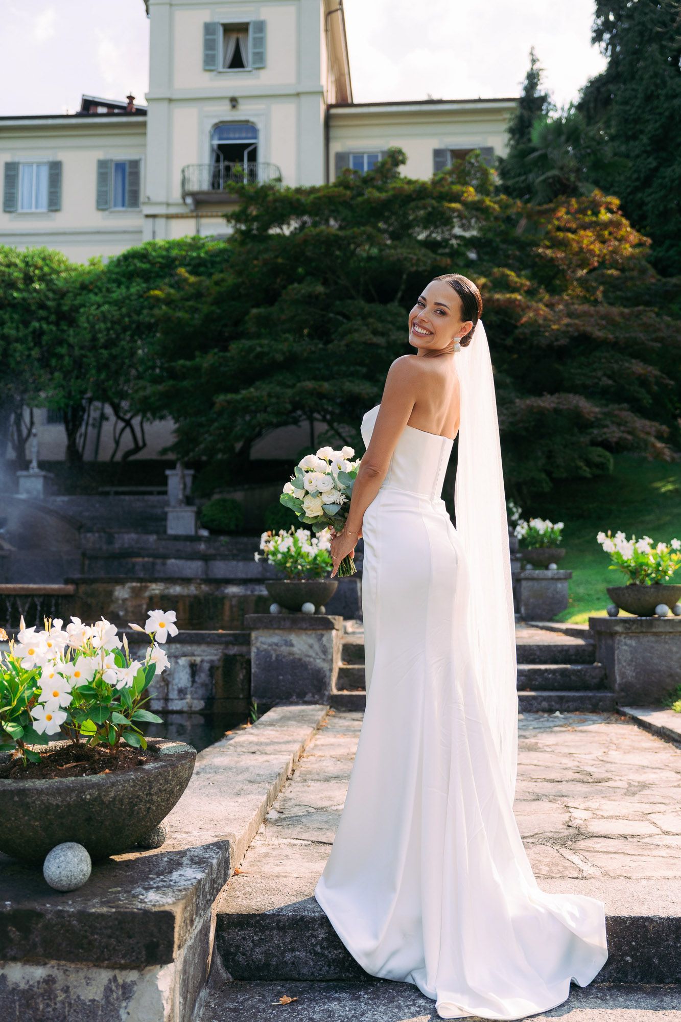 A bride in a flowing white wedding dress stands gracefully in a garden terrace with stone walls and flowering plants.