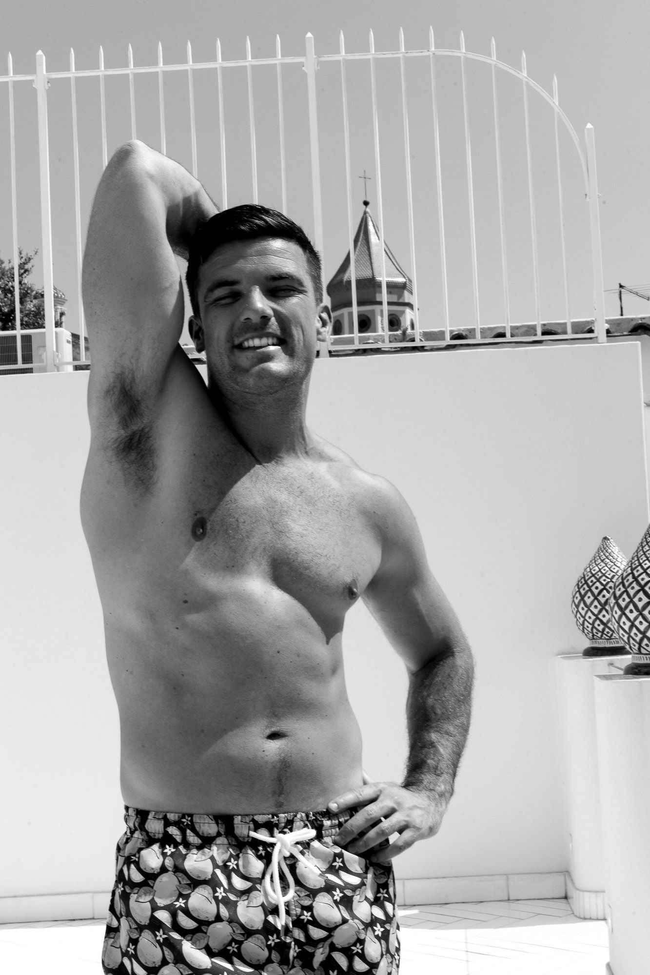 A person wearing patterned swim trunks poses shirtless in front of a white fence outdoors.