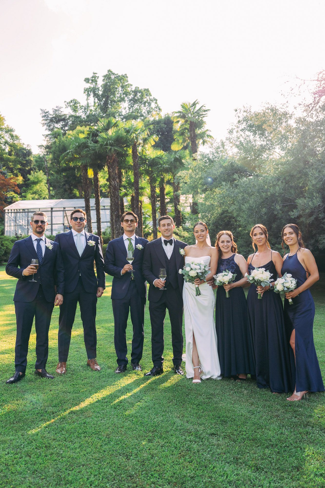 Wedding party in navy and white attire poses on green lawn during golden hour sunset.
