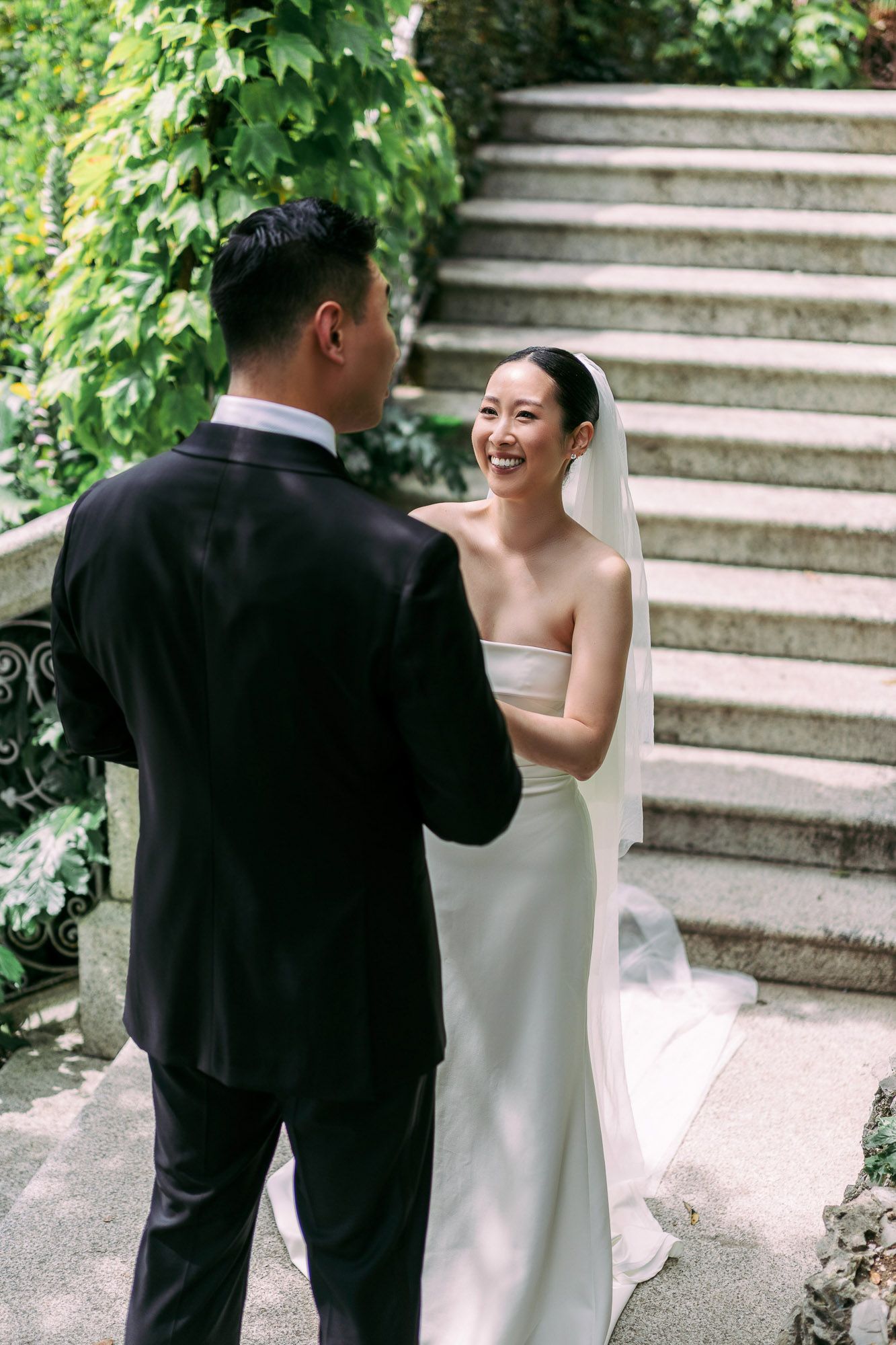 A bride and groom share a joyful first look moment on stone steps surrounded by lush green foliage.