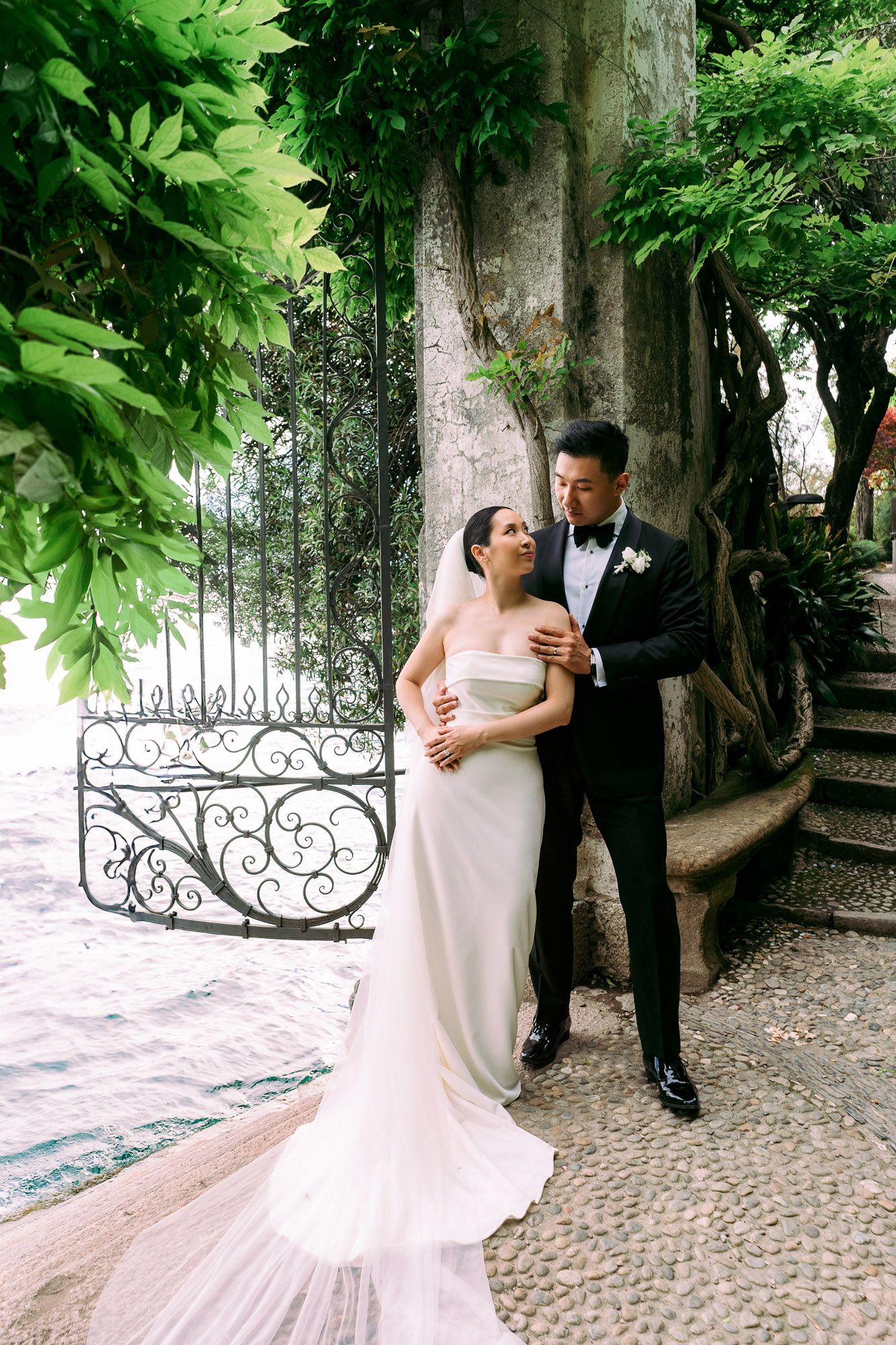 A romantic wedding couple poses together on a cobblestone path surrounded by lush greenery and ornate iron gates.