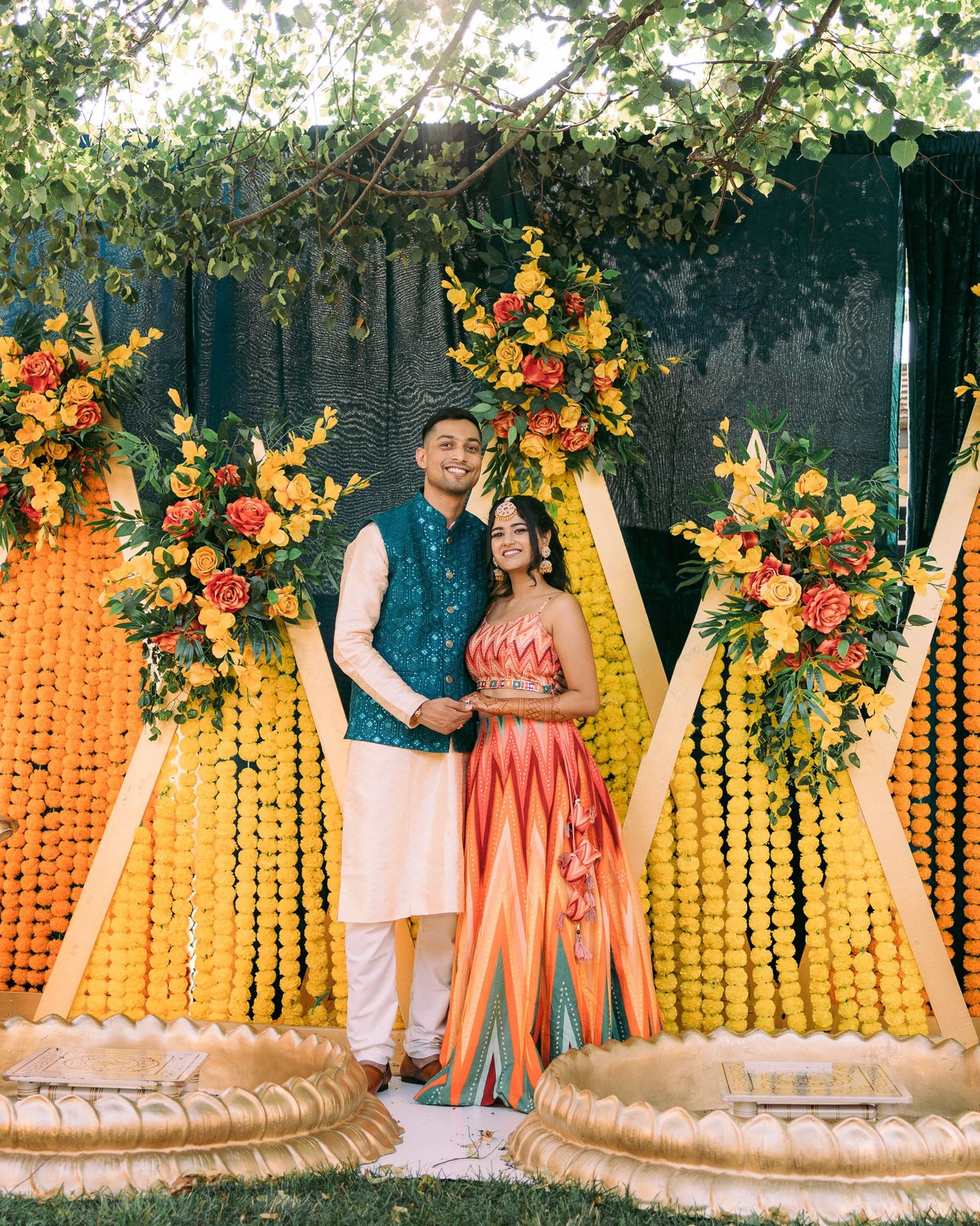 Outdoor wedding backdrop with marigold garlands and floral arrangements in orange and yellow against a natural garden setting.