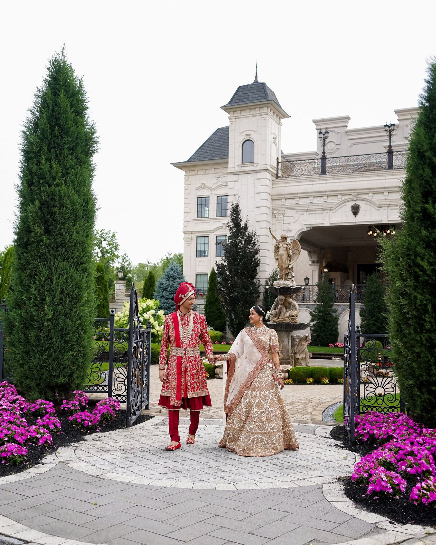Wedding couple poses in front of luxury mansion with vibrant pink flowers and cypress trees lining stone walkway.