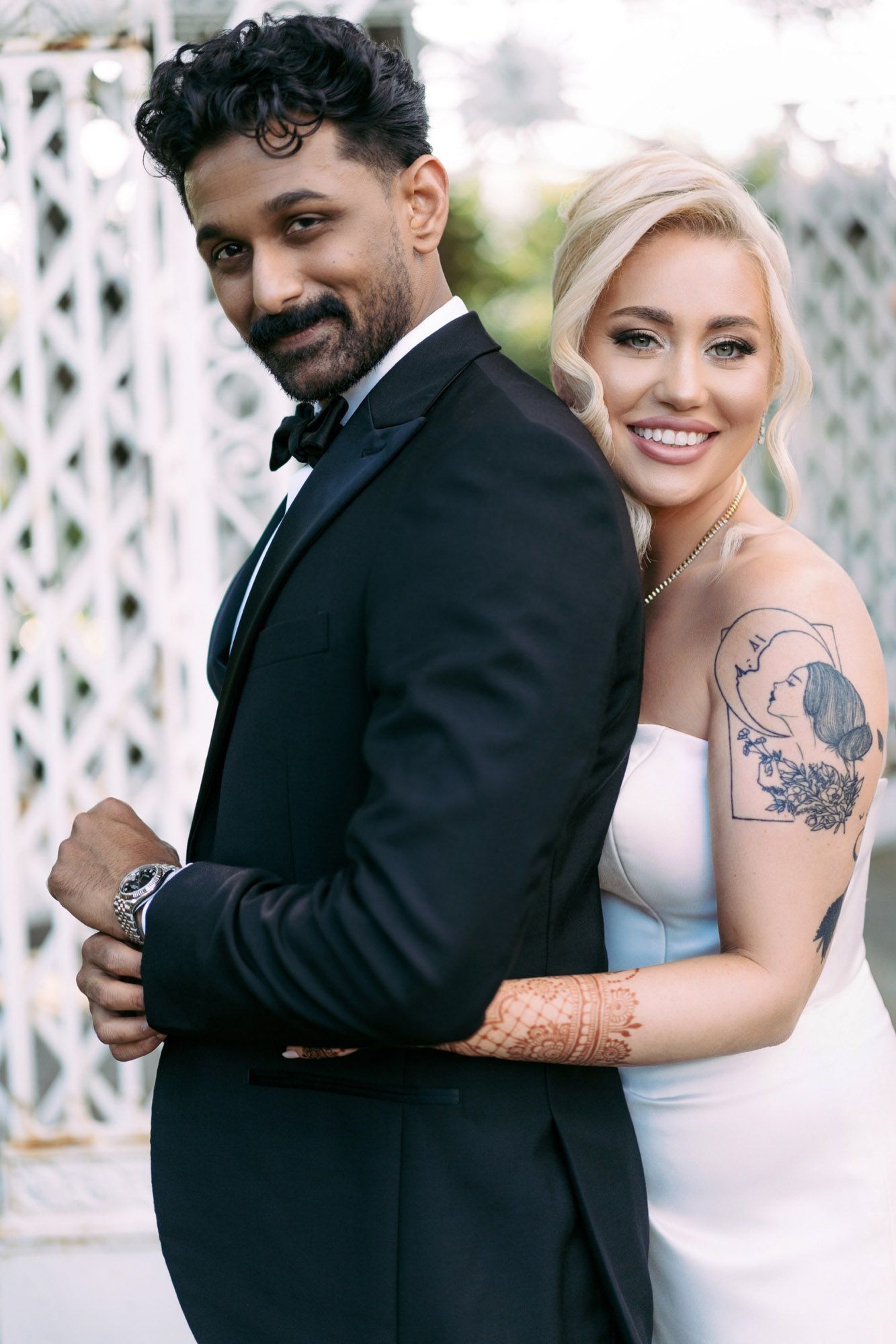 A happy couple in wedding attire poses together, with the bride wearing a white dress and showing a visible arm tattoo.