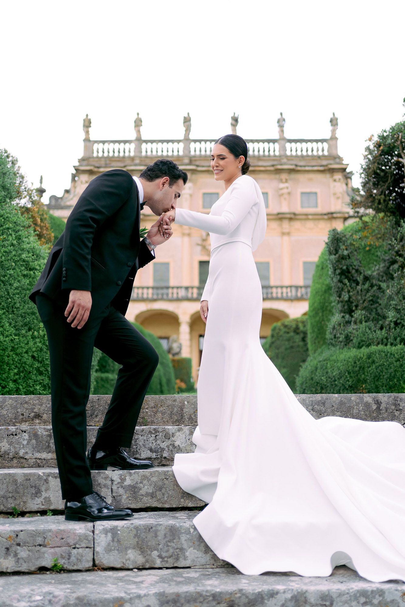 Elegant wedding couple on stone steps of historic villa with ornate balustrade and lush gardens in background.