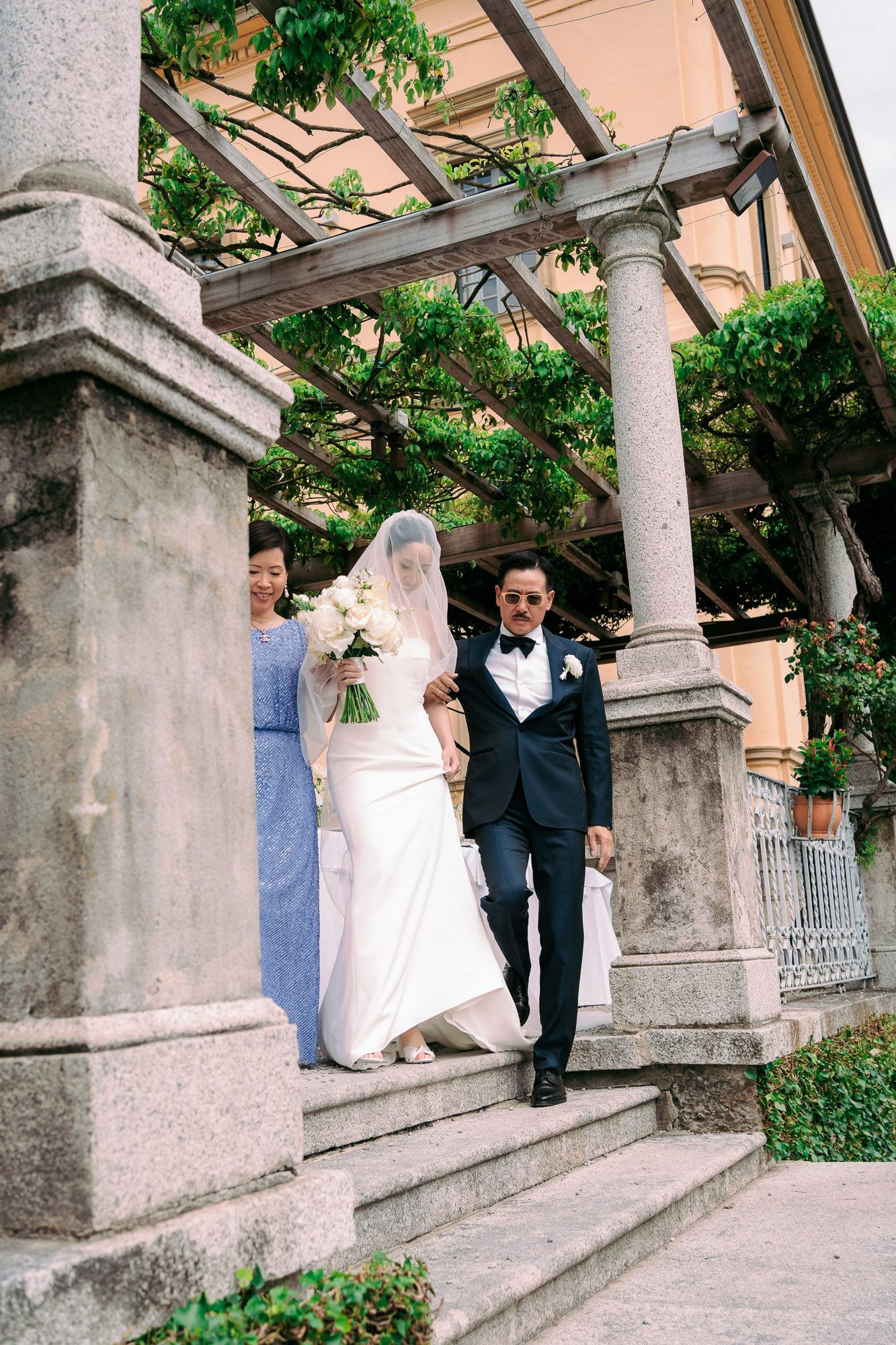 Wedding couple descends stone steps under an elegant columned pergola with climbing vines in a Mediterranean garden setting.