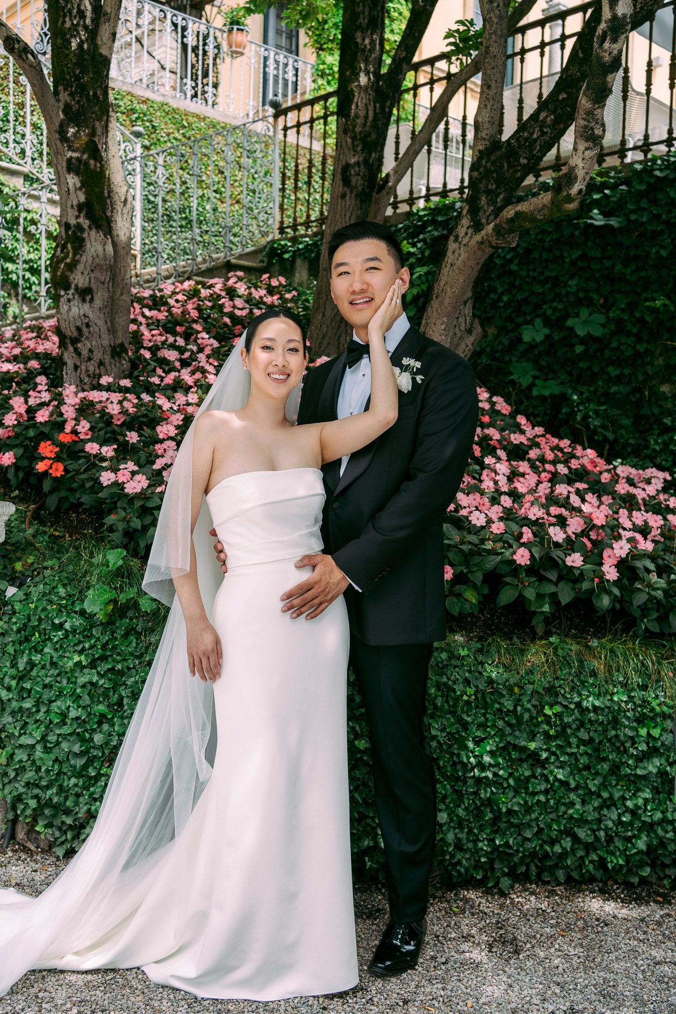 A couple shares joyful moments in a garden setting with pink flowers and greenery during their wedding photoshoot.