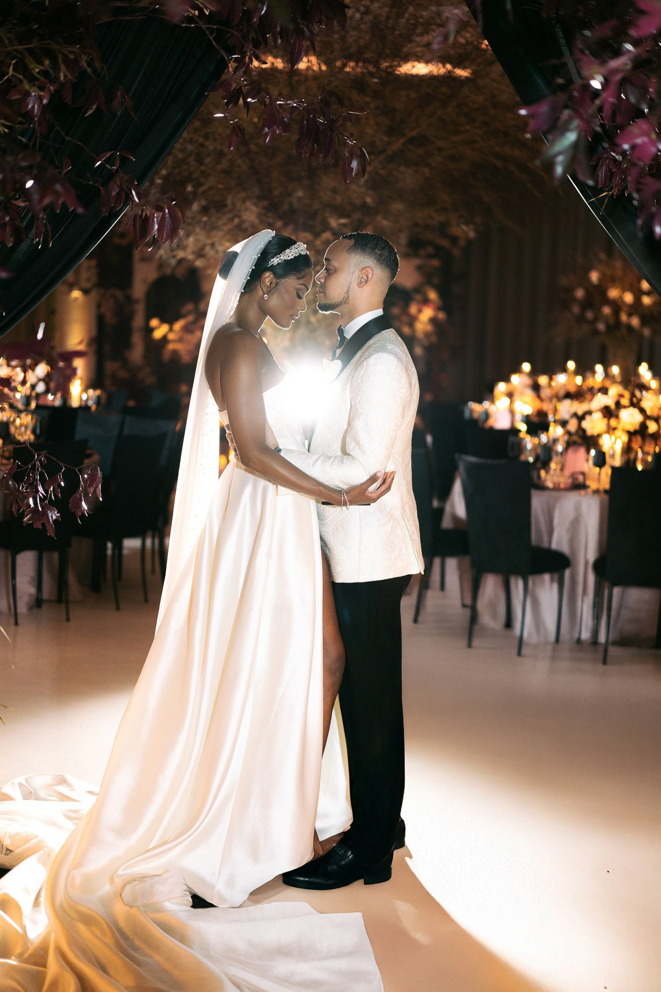 Romantic wedding couple embraces in an elegantly lit reception venue with flowing white dress and candlelit ambiance.