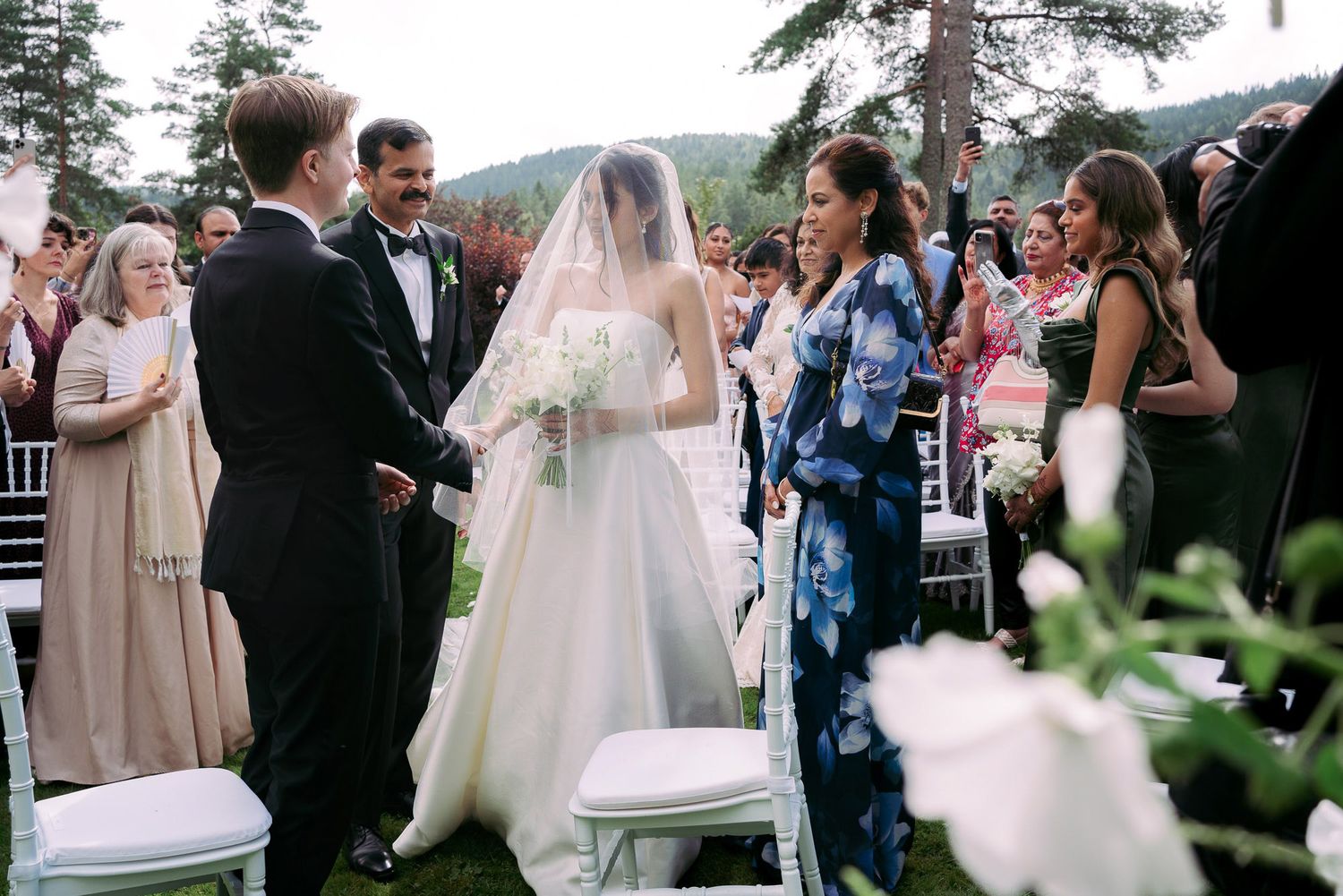 Wedding guests celebrate and gather outdoors during a summer ceremony with trees in the background.